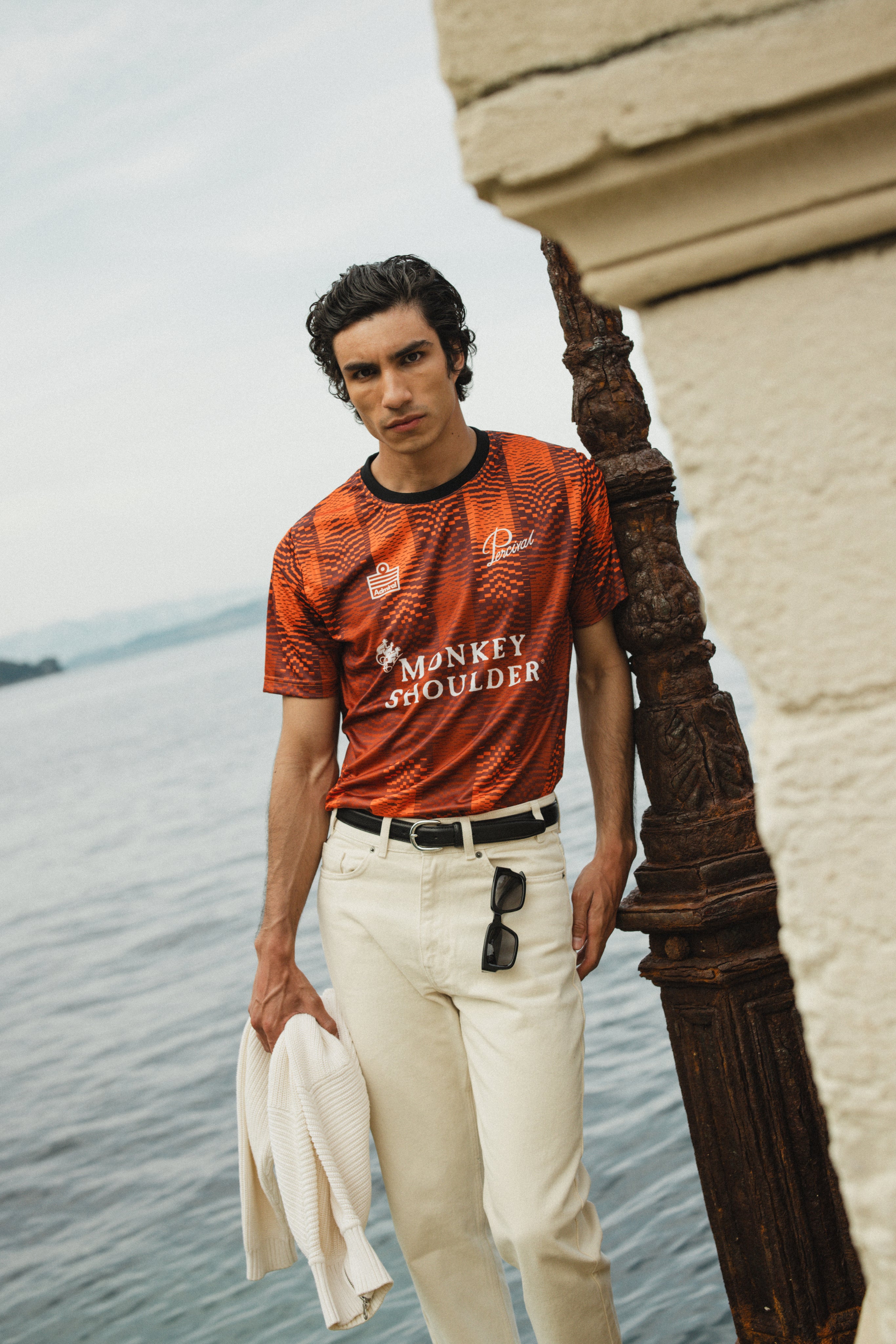 The Admiral x Percival x Monkey Shoulder orange pixel-distortion football shirt, styled with white trousers by the sea.