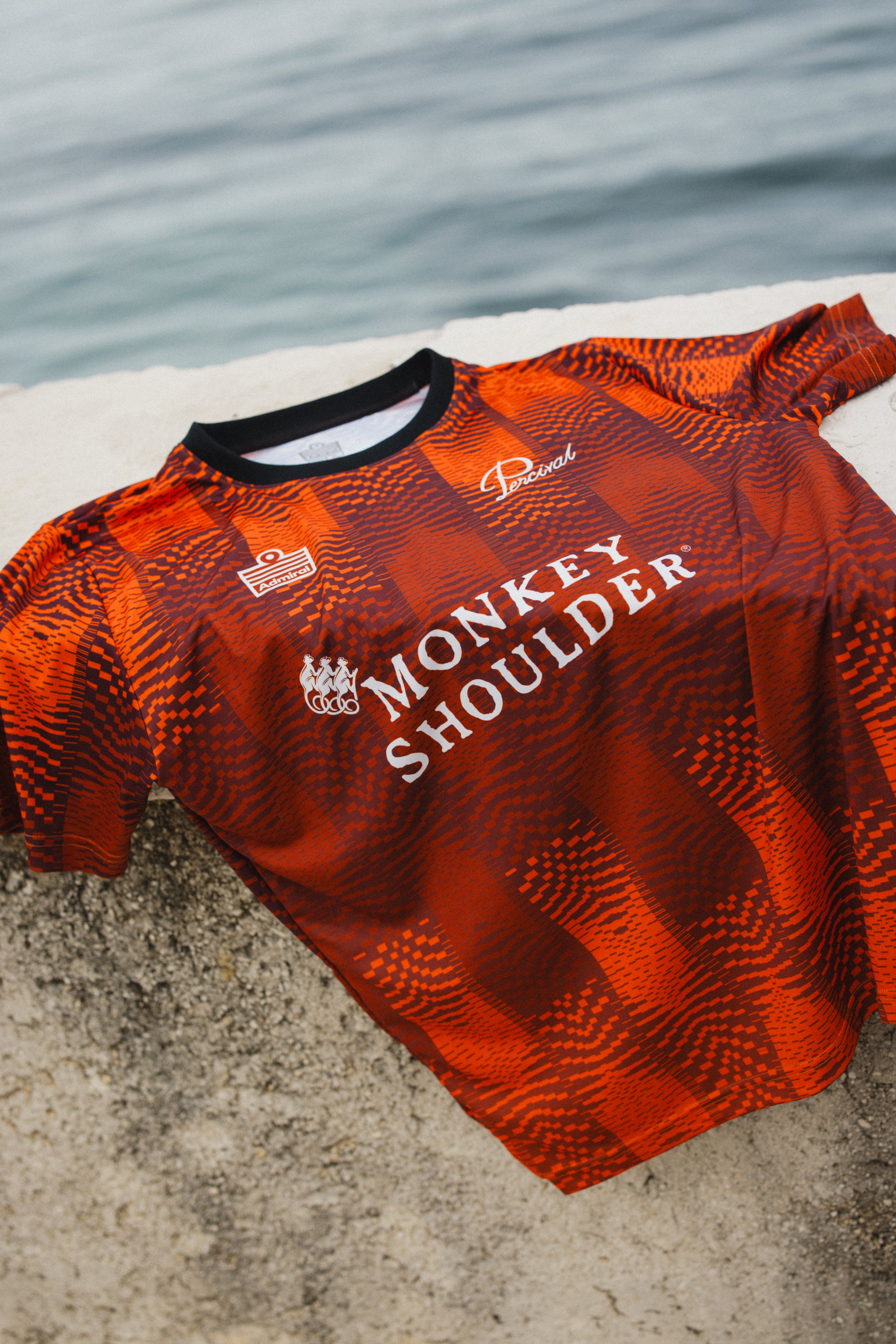 The orange Percival x Monkey Shoulder football shirt with a pixel-distortion stripe pattern, laid on a stone ledge.