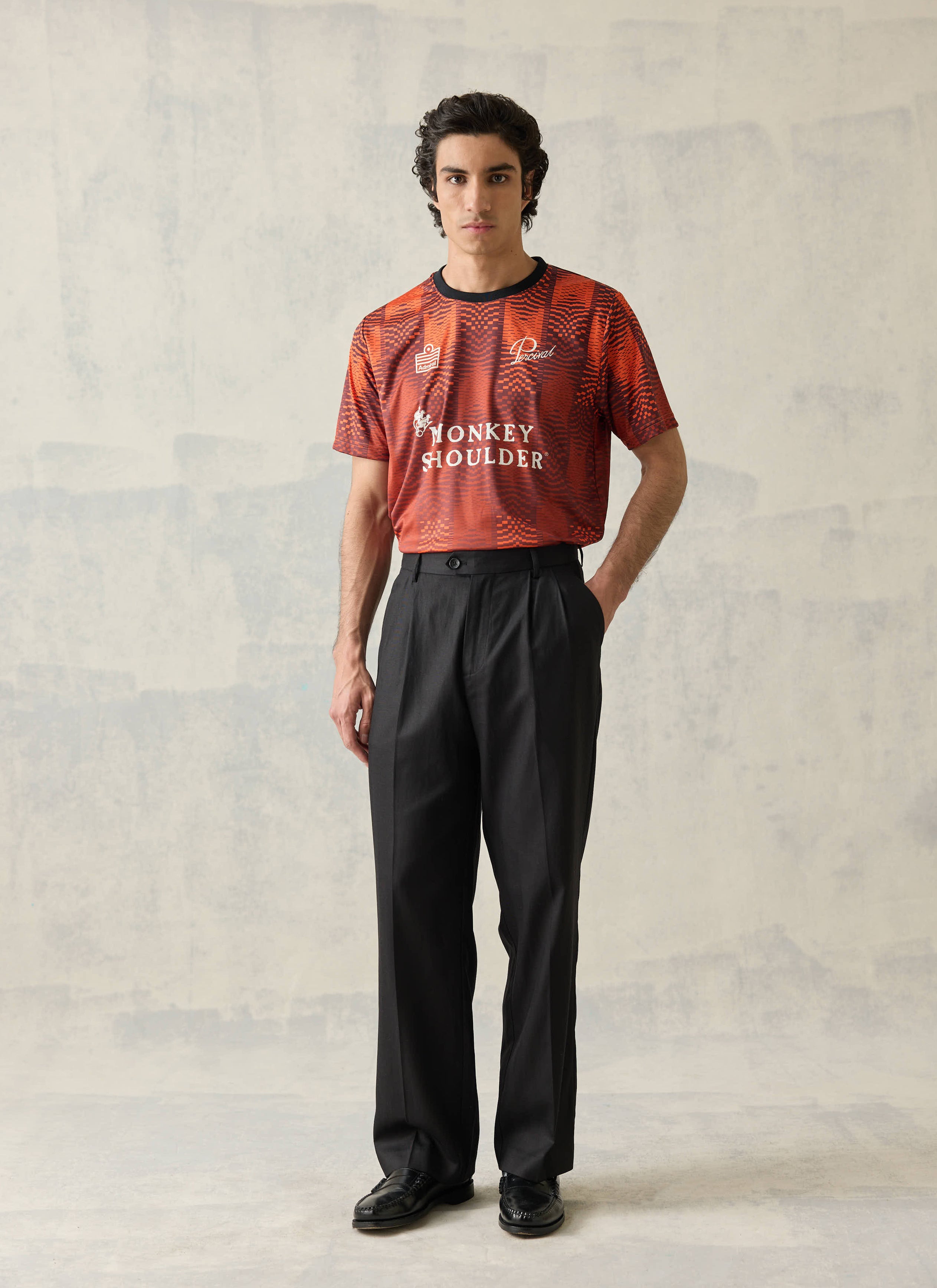 Orange Percival x Monkey Shoulder football shirt with pixel-distortion stripes, worn with black trousers.