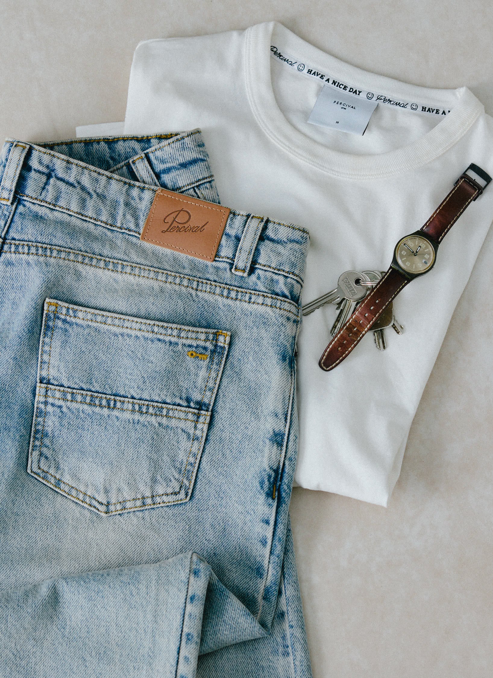 A still life of Percival stone wash straight leg denim jeans, a white Percival t-shirt, a watch, and a set of keys.