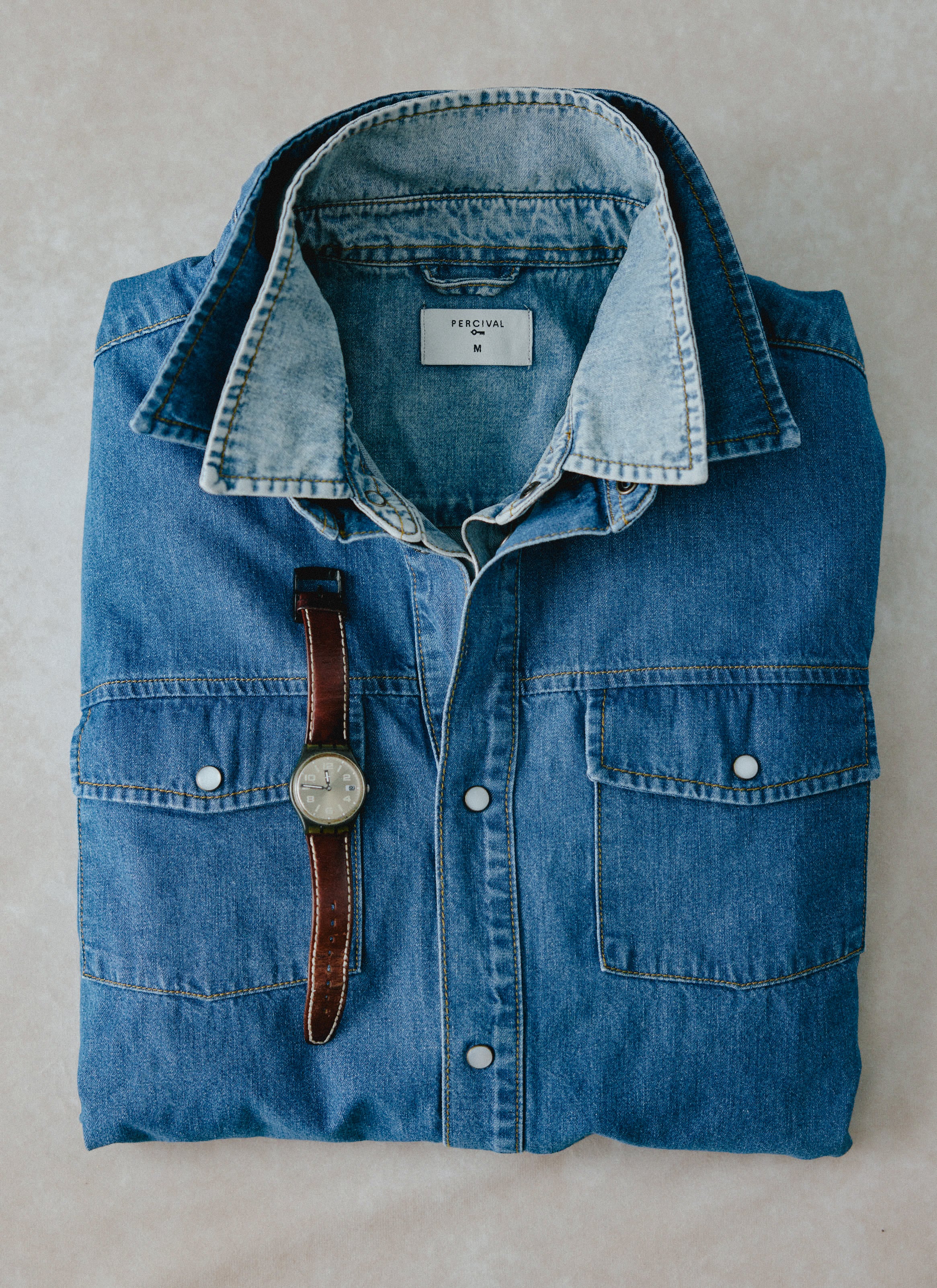 Still life of a folded Percival mid-wash blue denim western shirt with a brown leather strap watch on top.