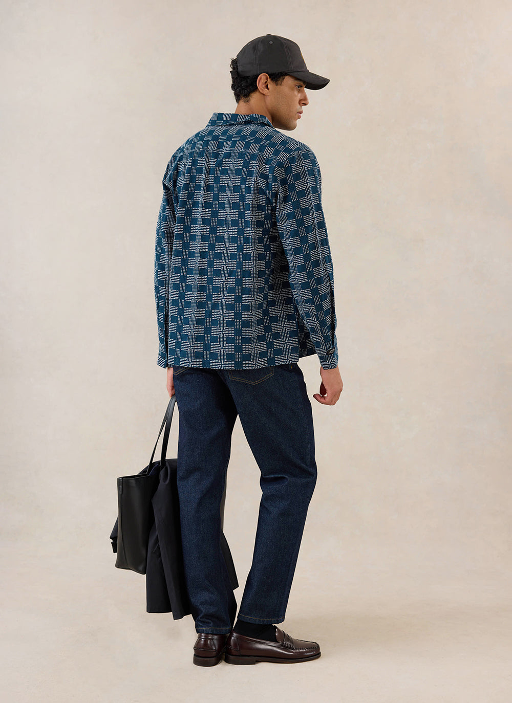 A model wears the navy Percival Nolan Jacquard Boxy Shirt from the back, styled with dark trousers, a cap and a bag.