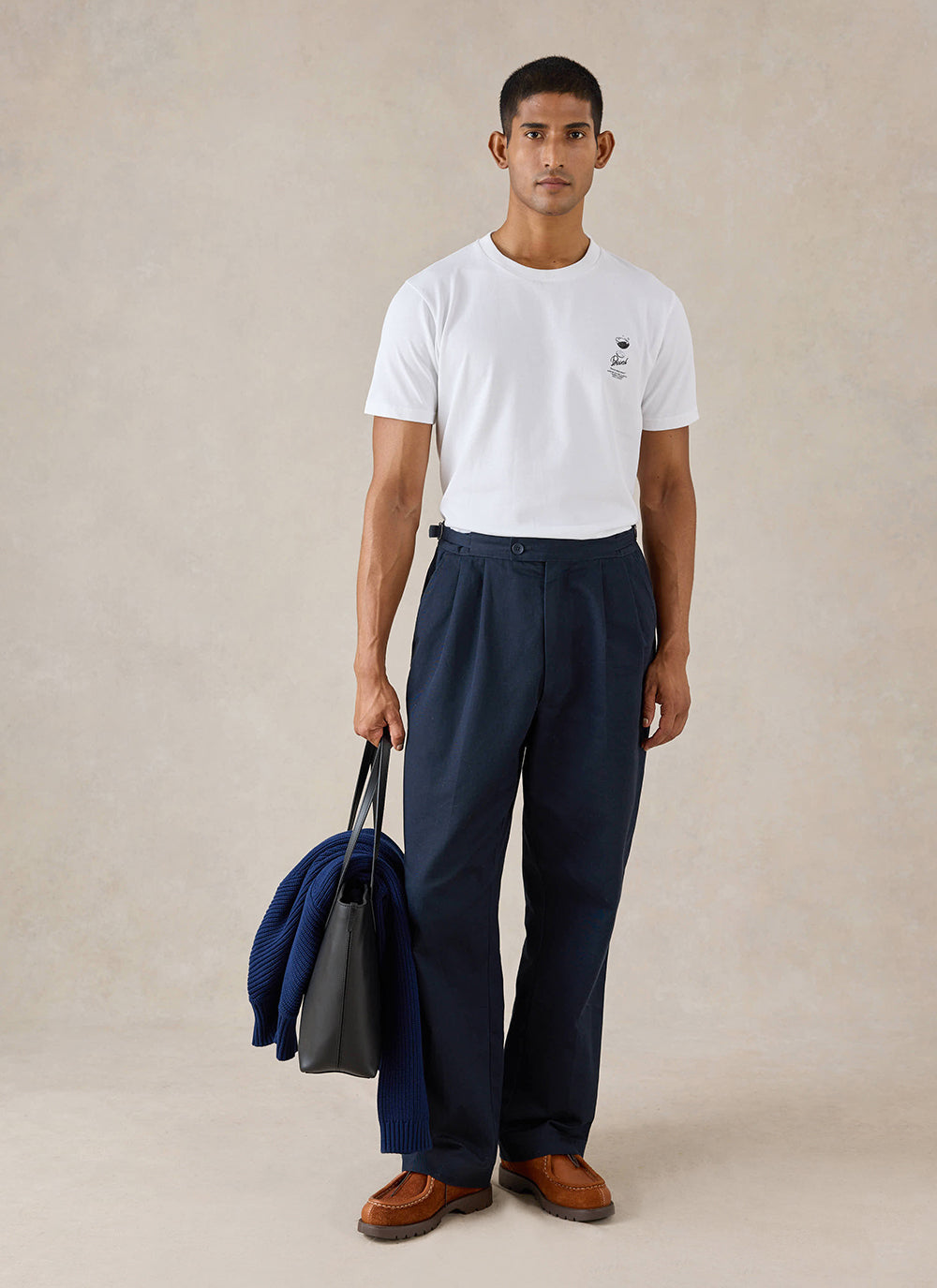 A model wears the white Percival Norman T-shirt, styled with navy trousers and a black leather tote bag in a studio.