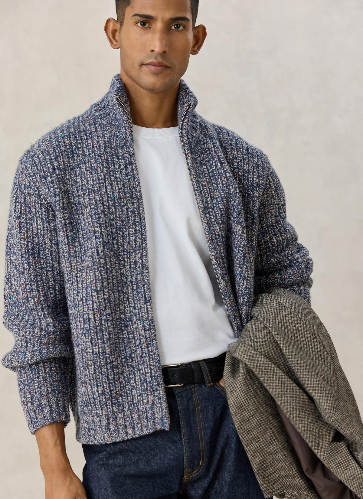 A model wearing the Percival blue fleck alpaca-wool zip cardigan over a white t-shirt and jeans, holding a tweed jacket.