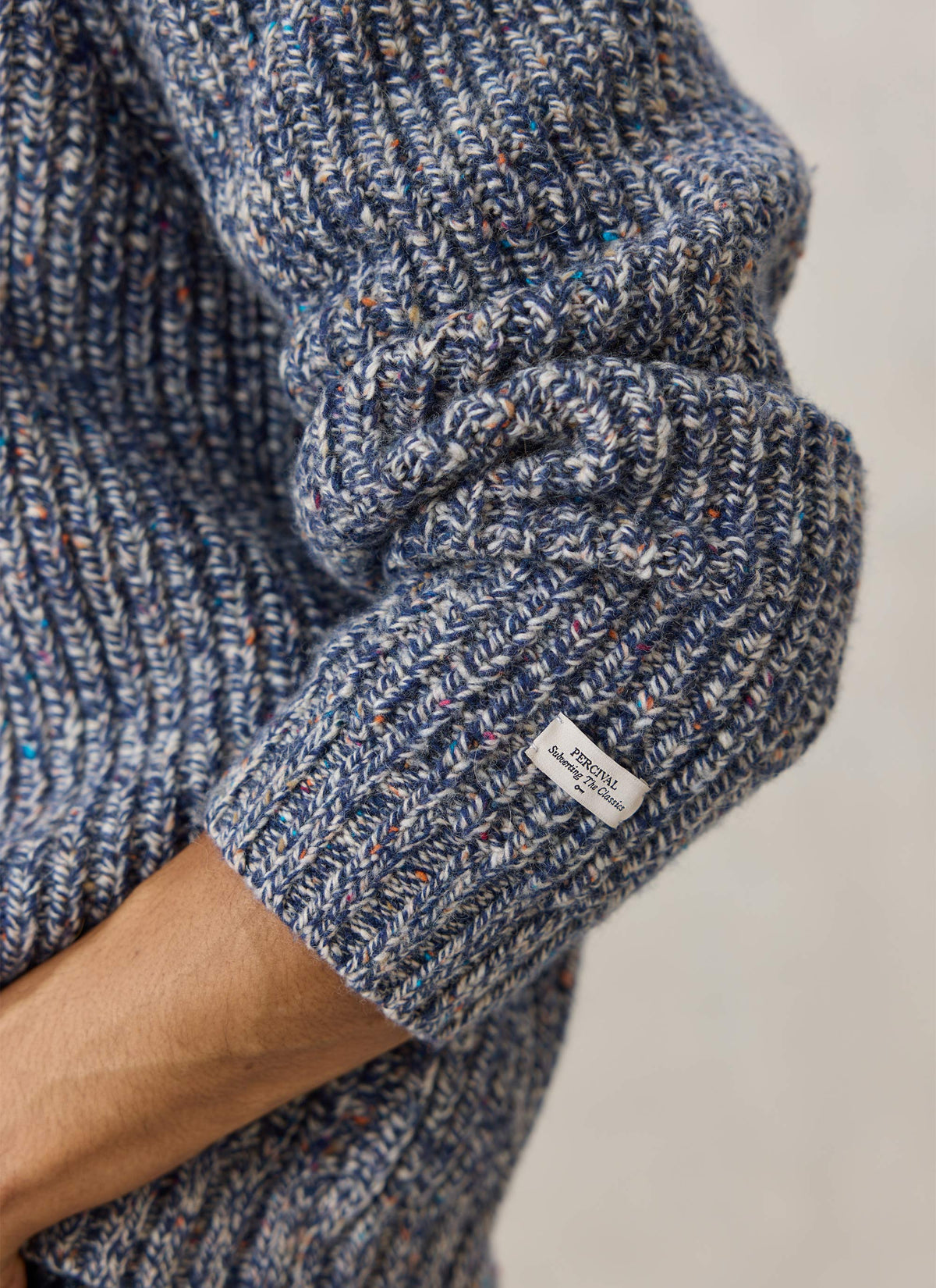 A close-up of the Percival Norton blue fleck alpaca-wool zip cardigan sleeve showing the ribbed knit and woven brand label.
