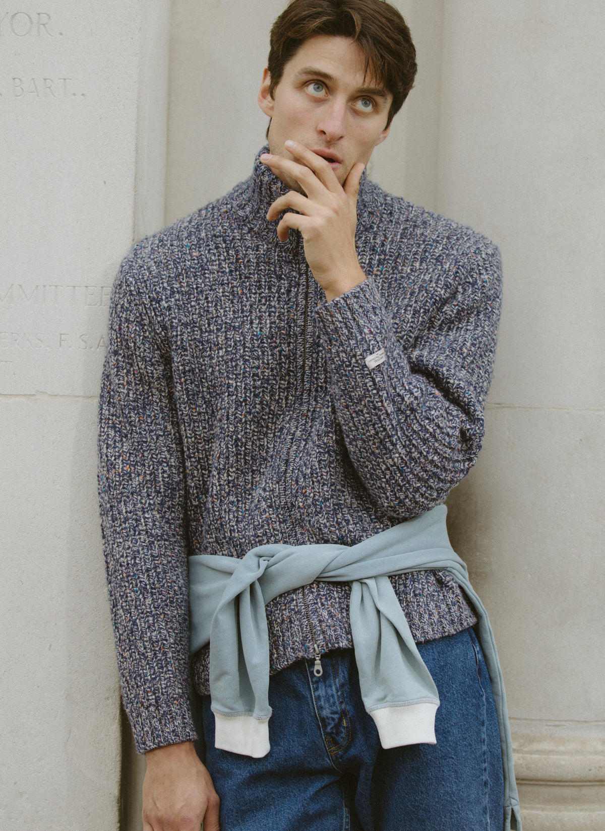 A model wearing the Percival blue fleck fisherman zip cardigan in alpaca wool with jeans, leaning against a wall.