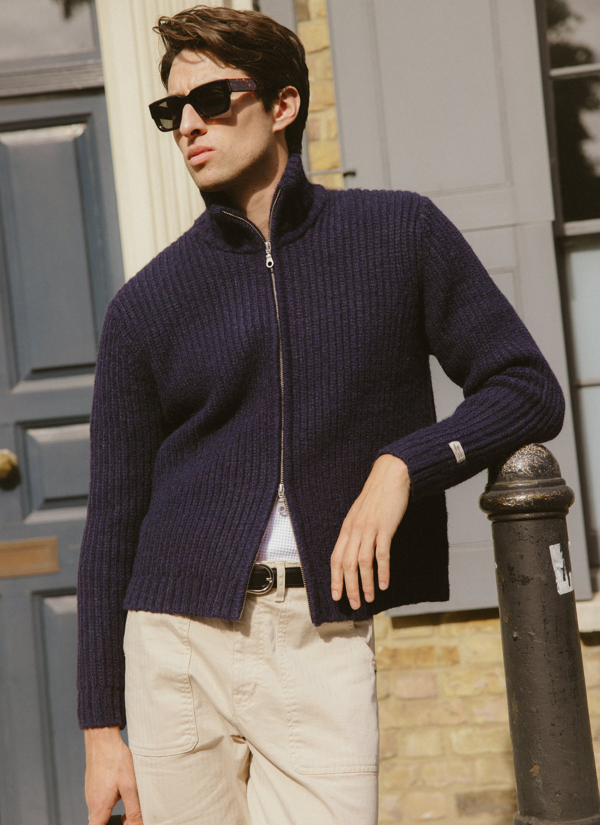 A model wearing the Percival navy ribbed fisherman zip cardigan in an alpaca wool blend outdoors against a building backdrop.