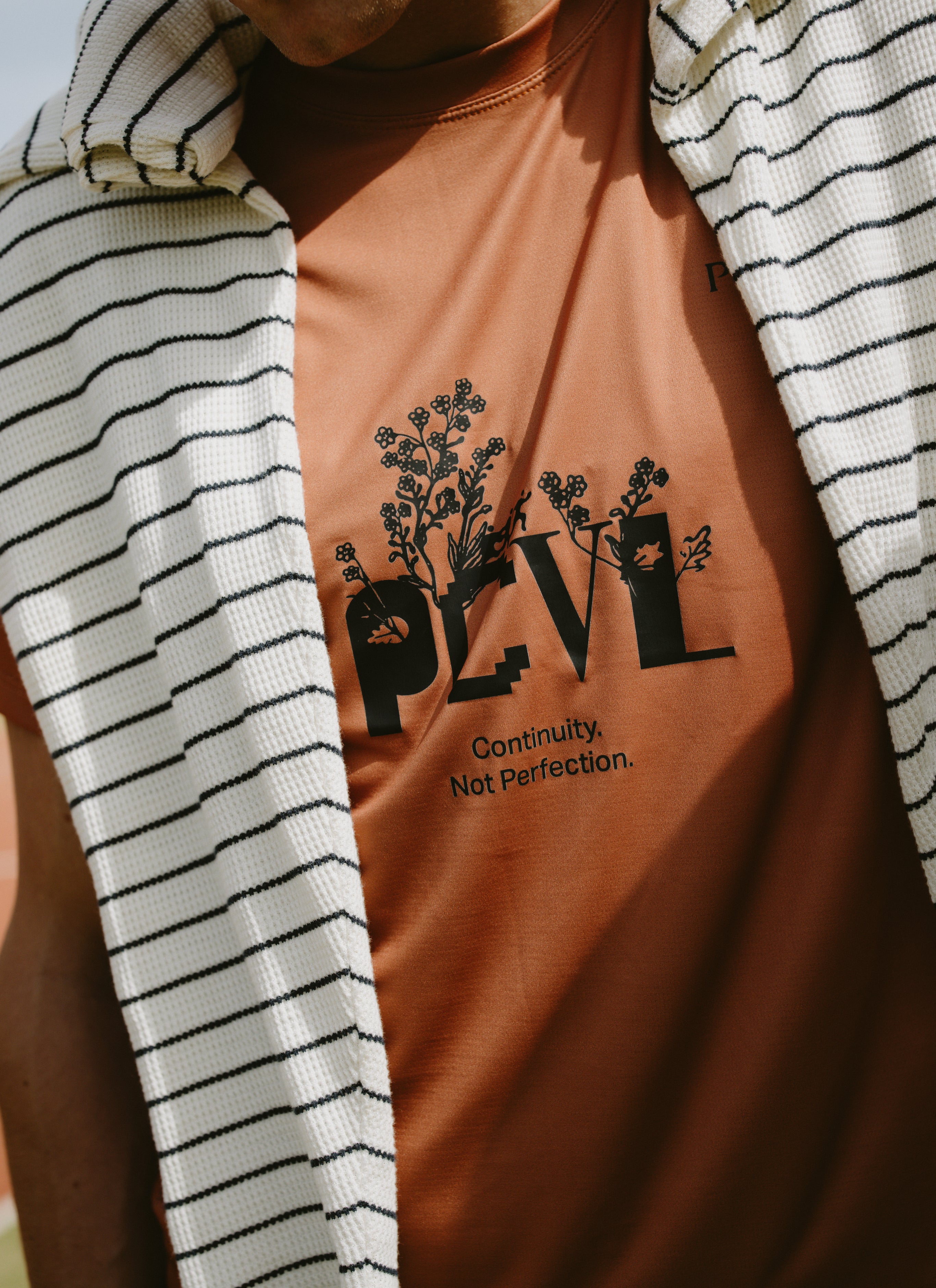 Percival rust orange performance top featuring floral PCVL graphics and text, layered with a striped knit cardigan.