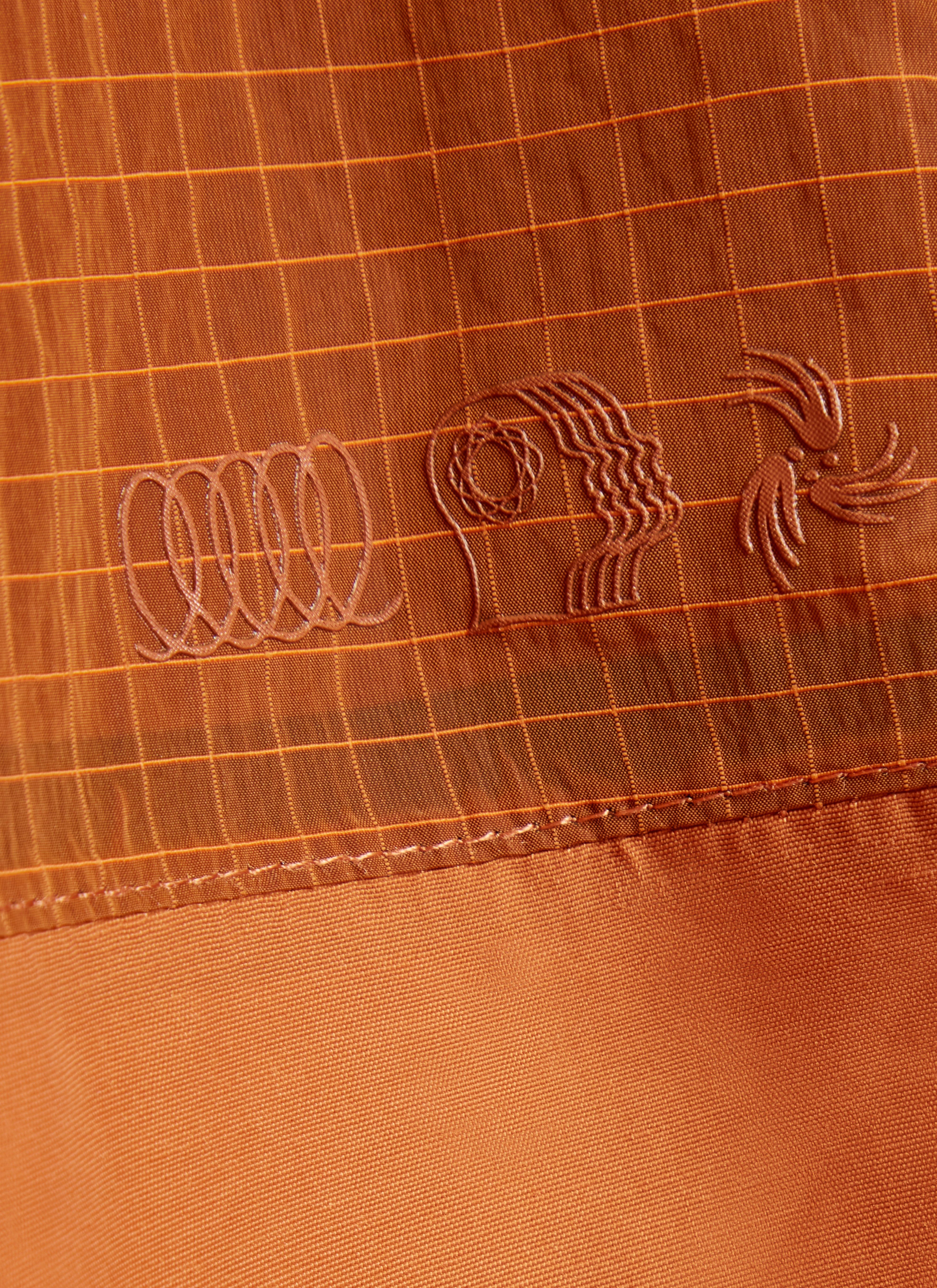 Close-up of the tonal PCVL logo on the rust water-resistant jacket from Percival, showing the technical grid fabric.
