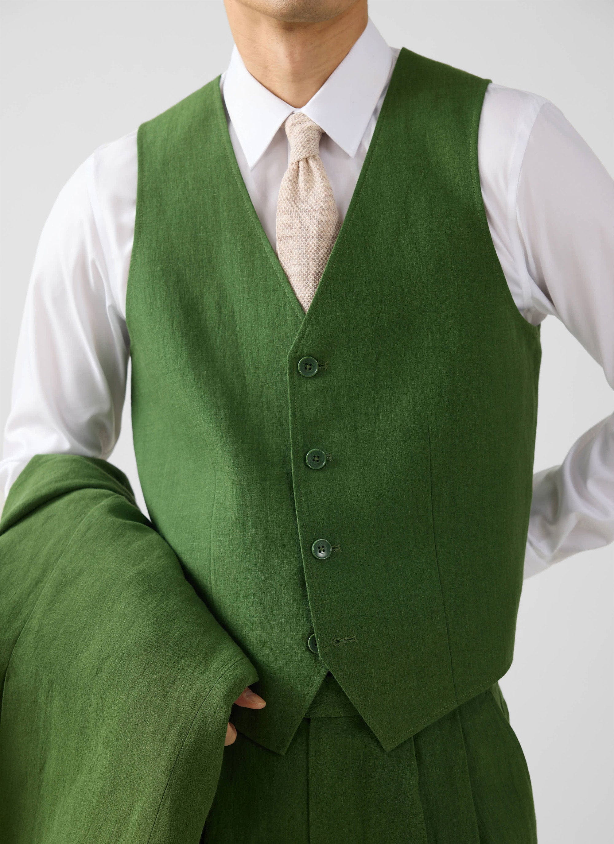 A model wears the Percival Linen Waistcoat in Forest over a white shirt and cream tie, holding the matching blazer.