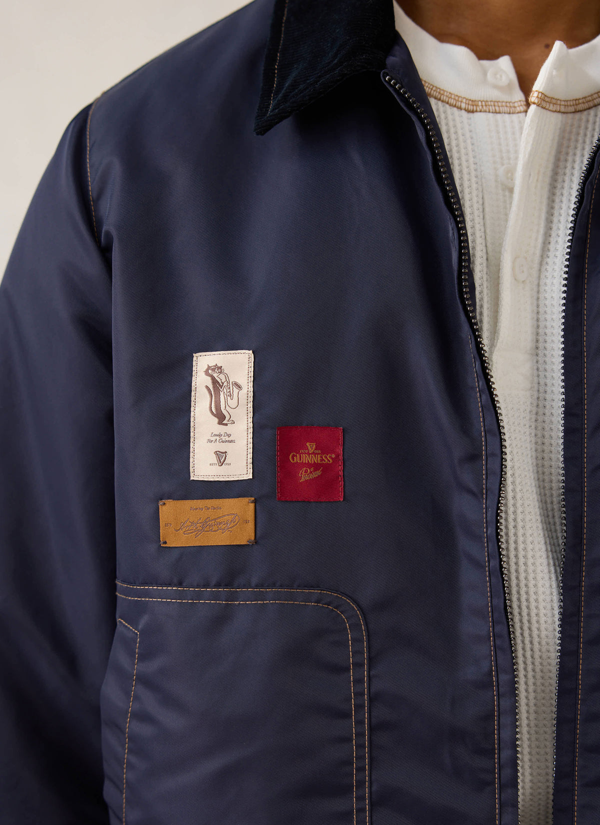 Close-up of the Guinness x Percival navy nylon jacket showing embroidered patches, contrast stitching, and cord collar.