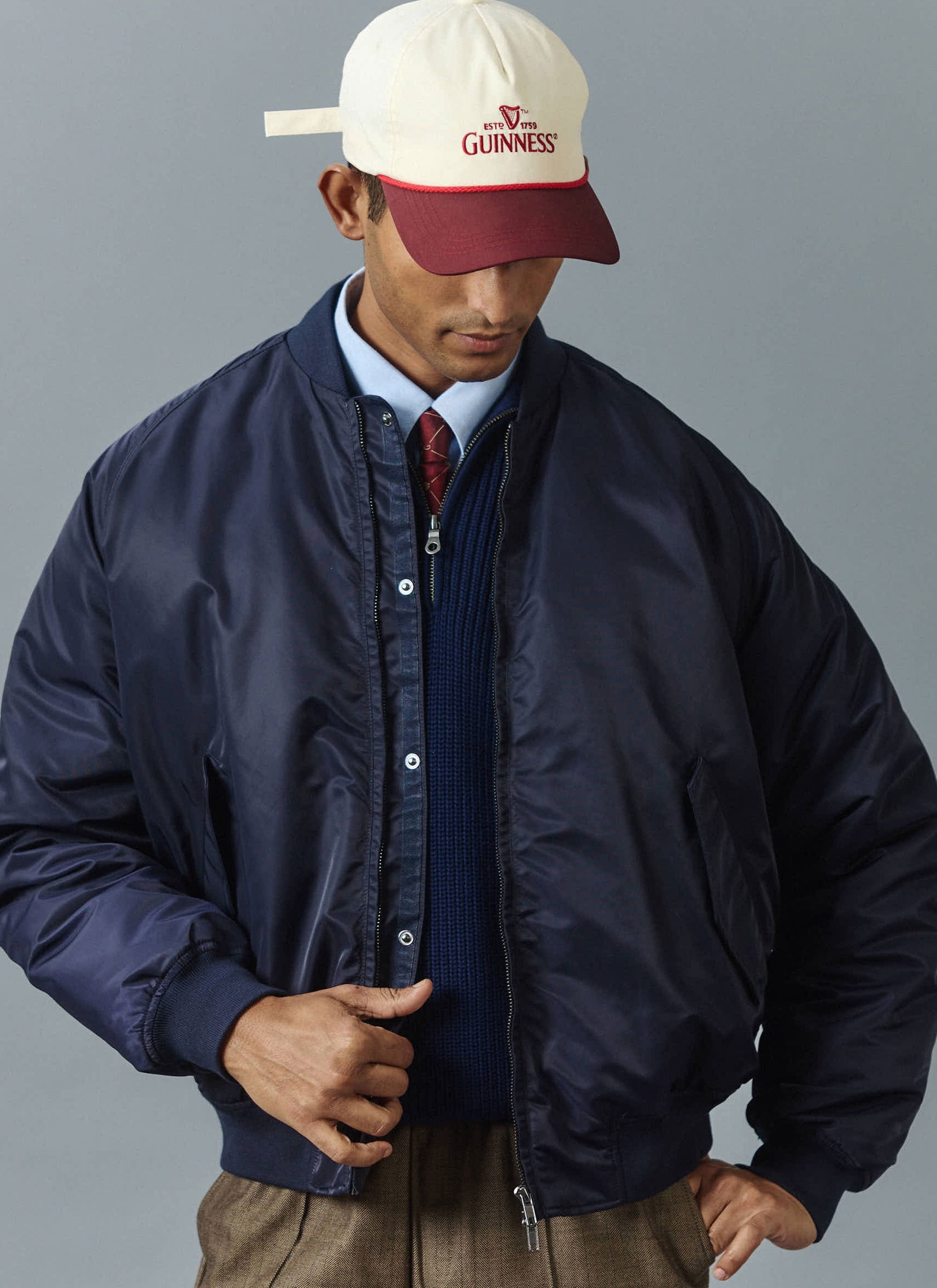 A man models the Percival midnight navy nylon bomber jacket, worn open over a blue jumper, shirt, and patterned red tie.