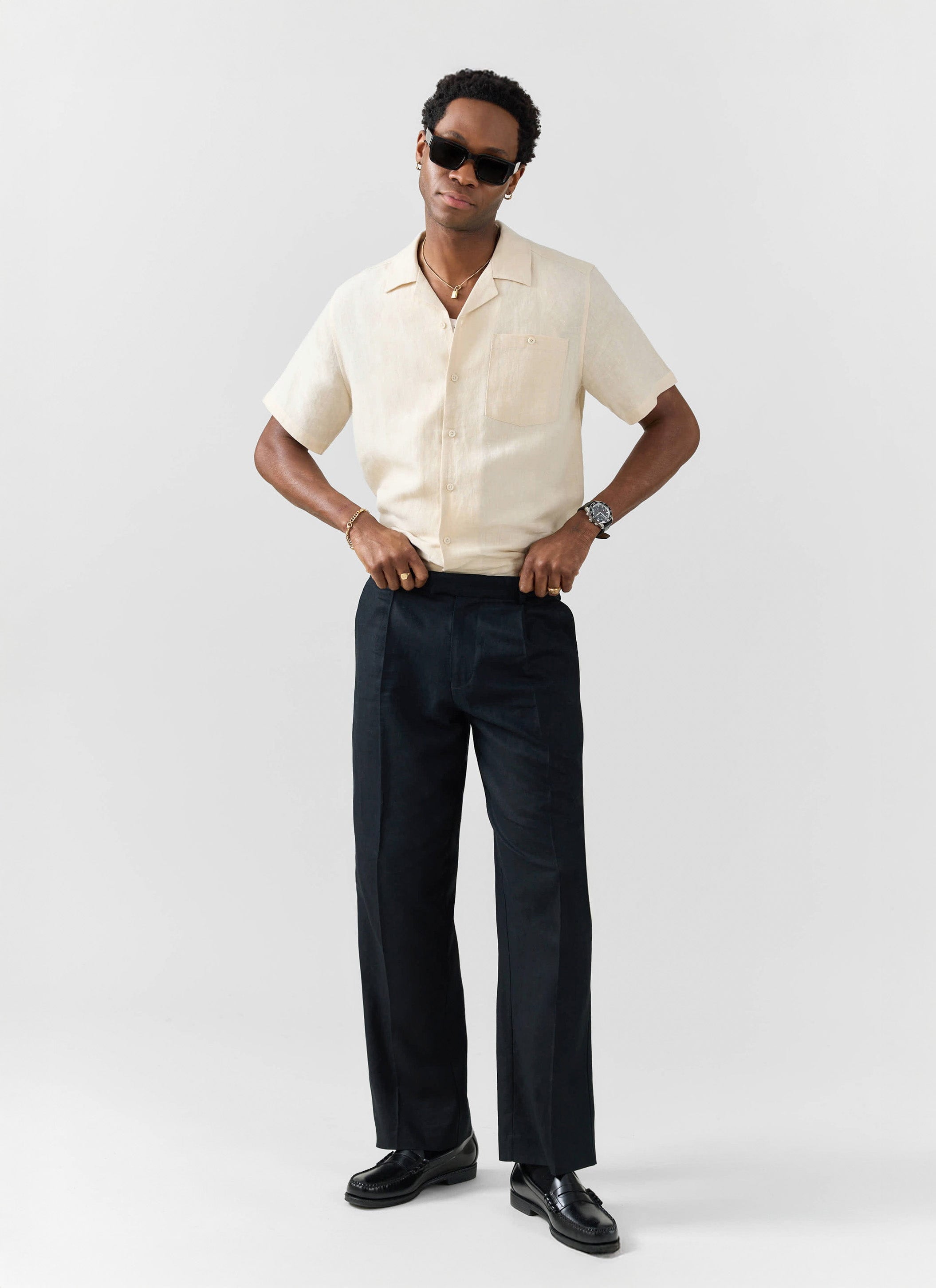 Model wears Percival cream linen short sleeve cuban shirt styled with black trousers, loafers and sunglasses.