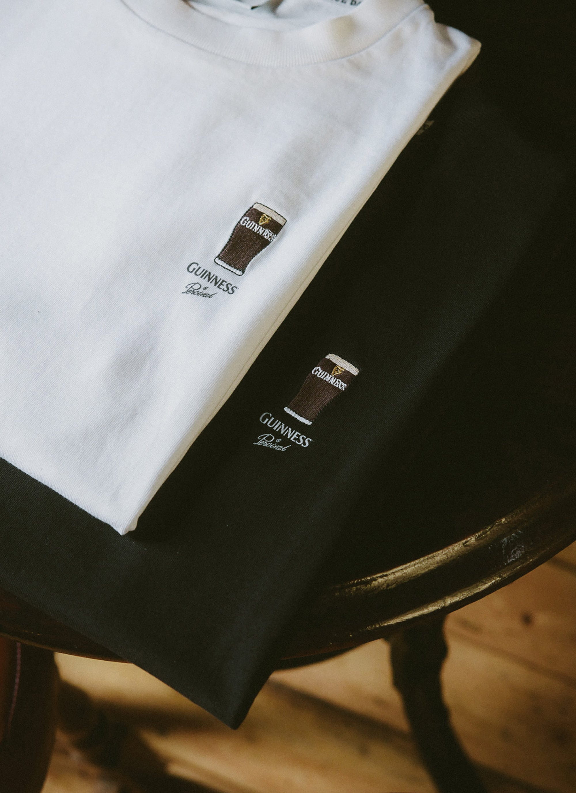 The black and white Percival x Guinness Stout T-shirts folded on a wooden table to showcase the pint embroidery.
