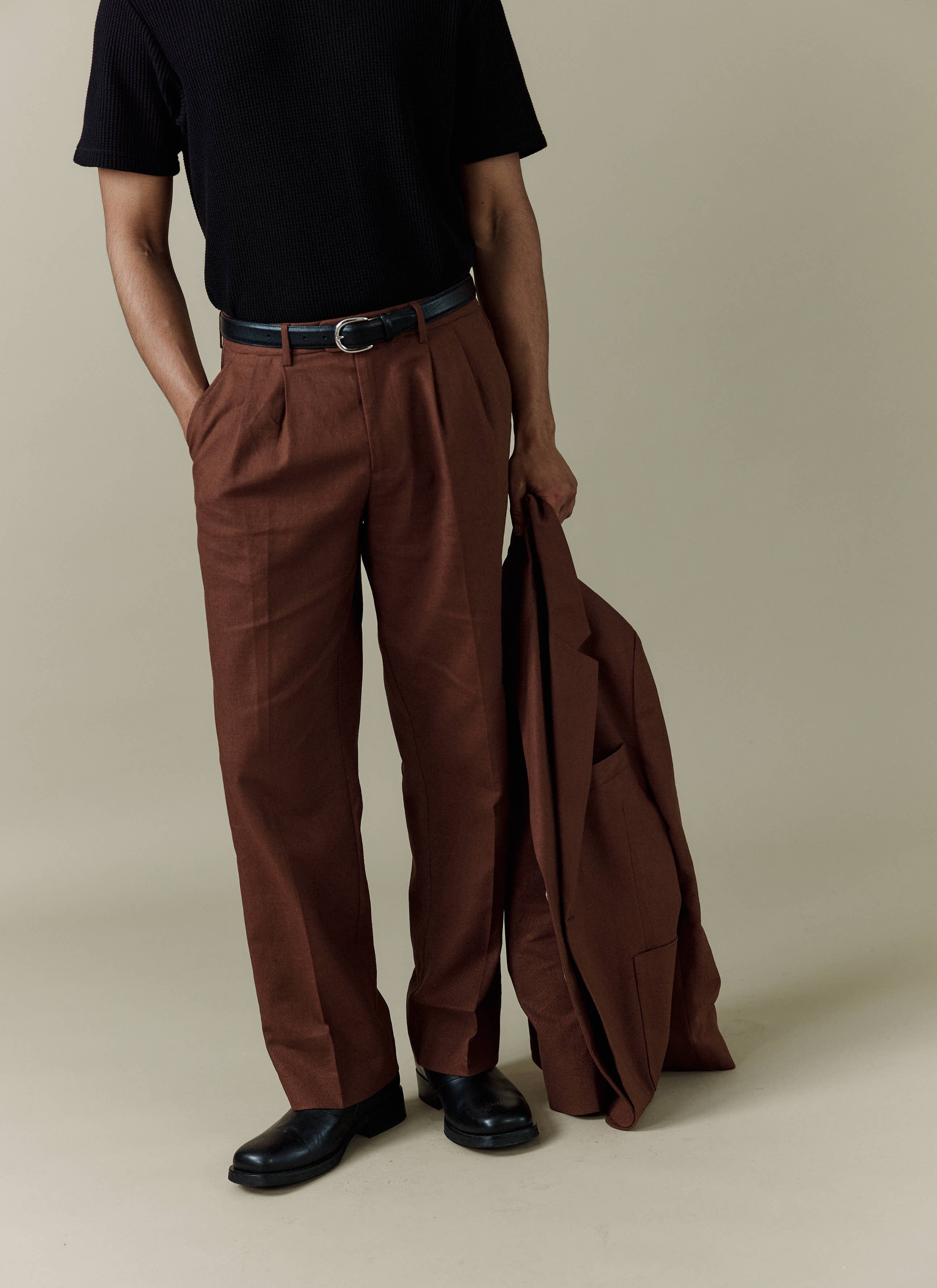 A model wears the Percival brown linen Pleated Tailored Trousers styled with a black t-shirt and holding the matching jacket.