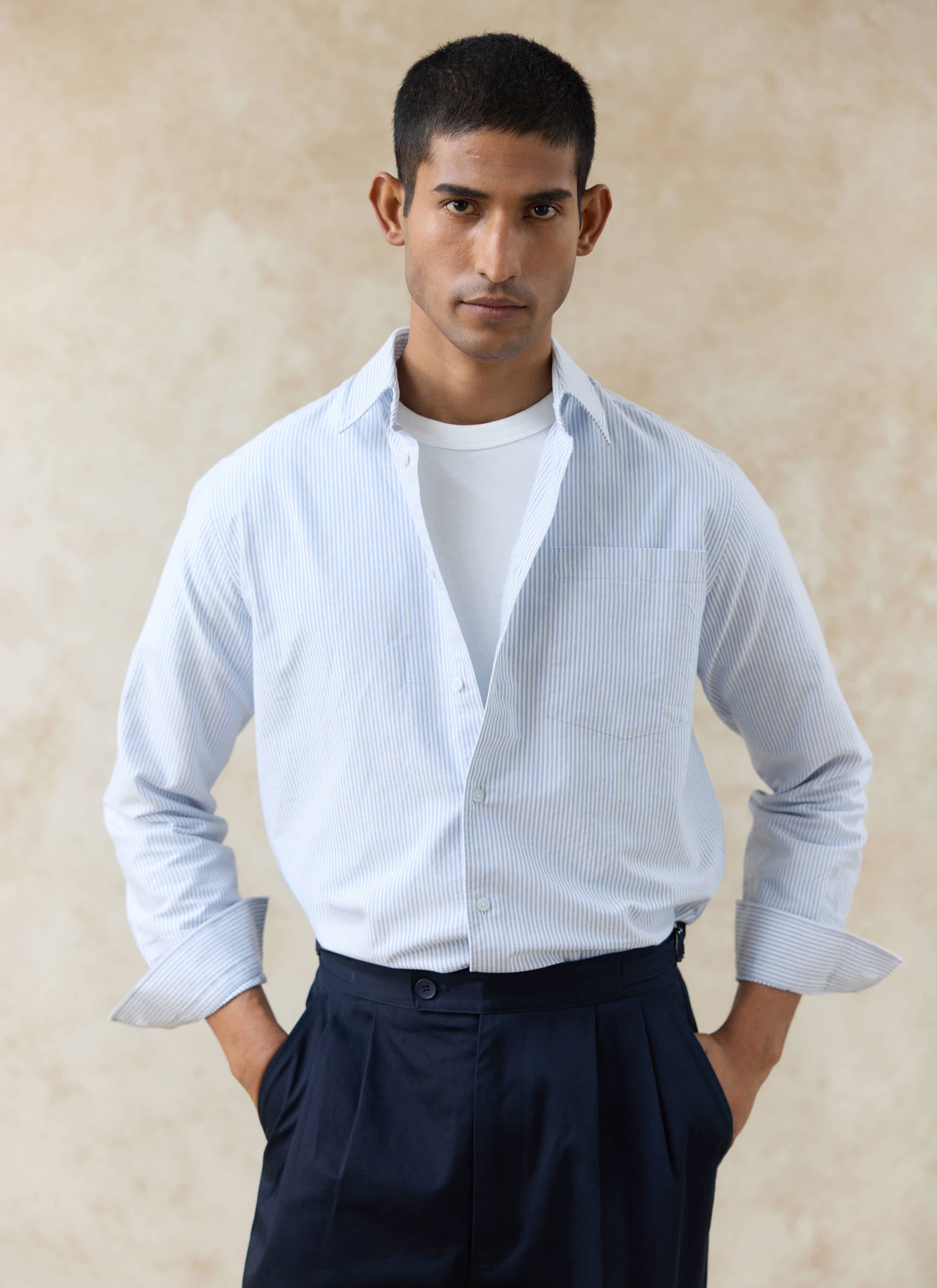 Model wears the Percival Striped Oxford Regular Shirt in blue cotton, layered over a white tee with navy trousers.