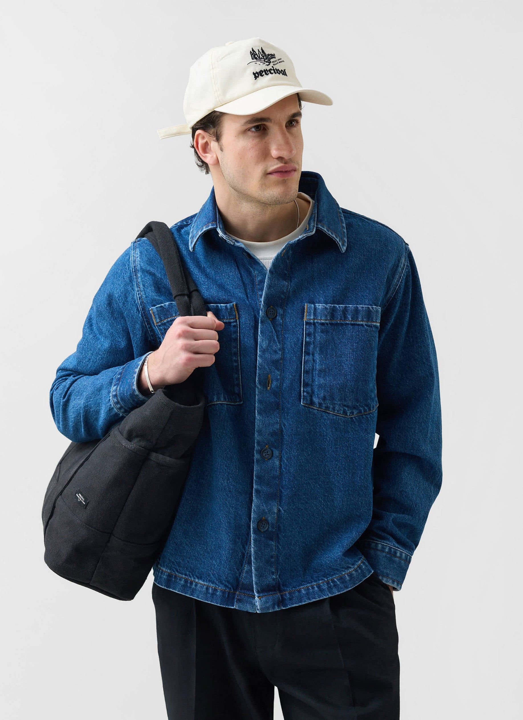 A model wears the Percival Denim Heavyweight Overshirt in mid-wash blue, styled with a cap and black shoulder bag.
