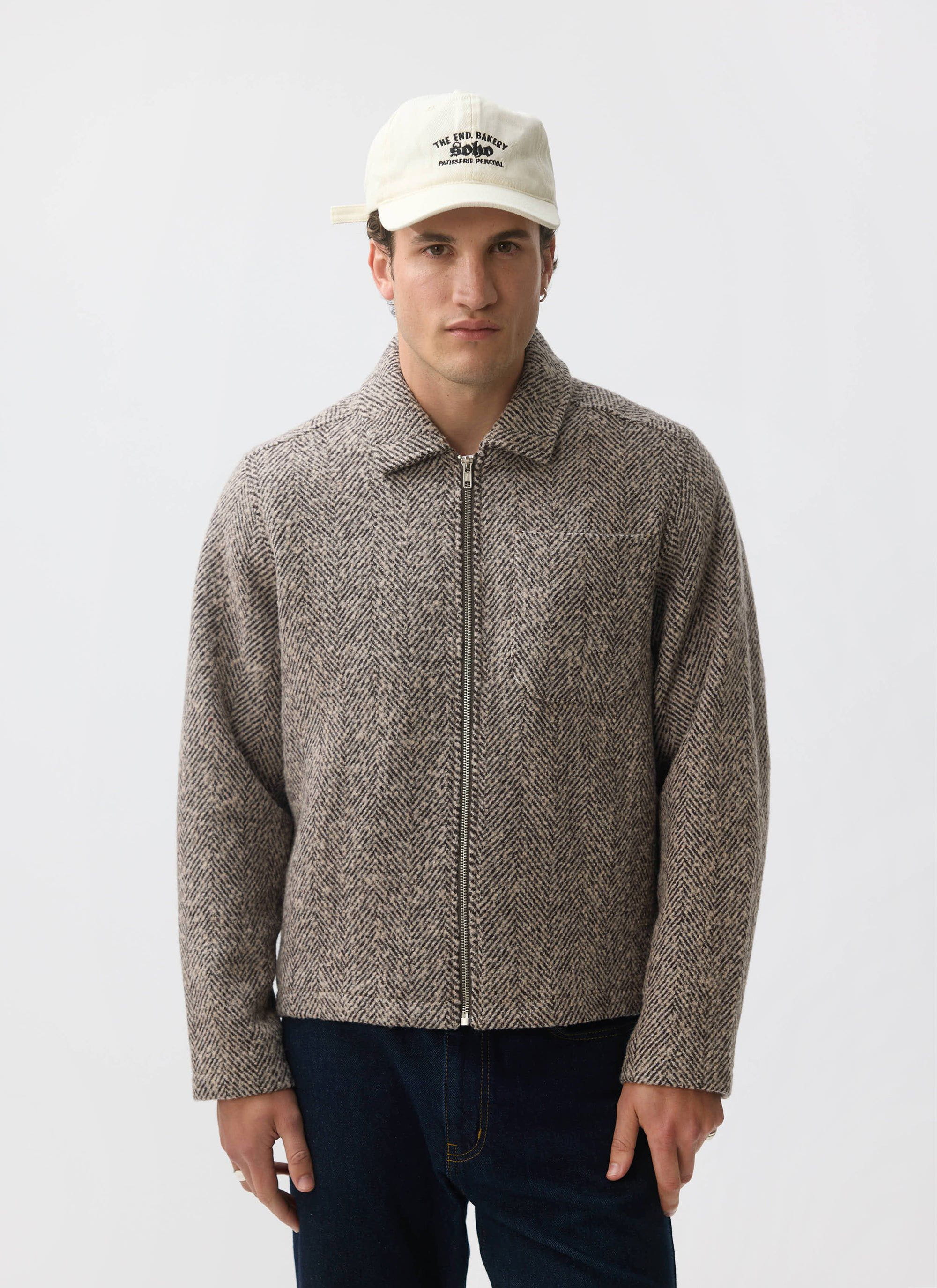 A model wears the Percival Herringbone Zip Jacket in ecru wool, paired with dark jeans and a white cap.