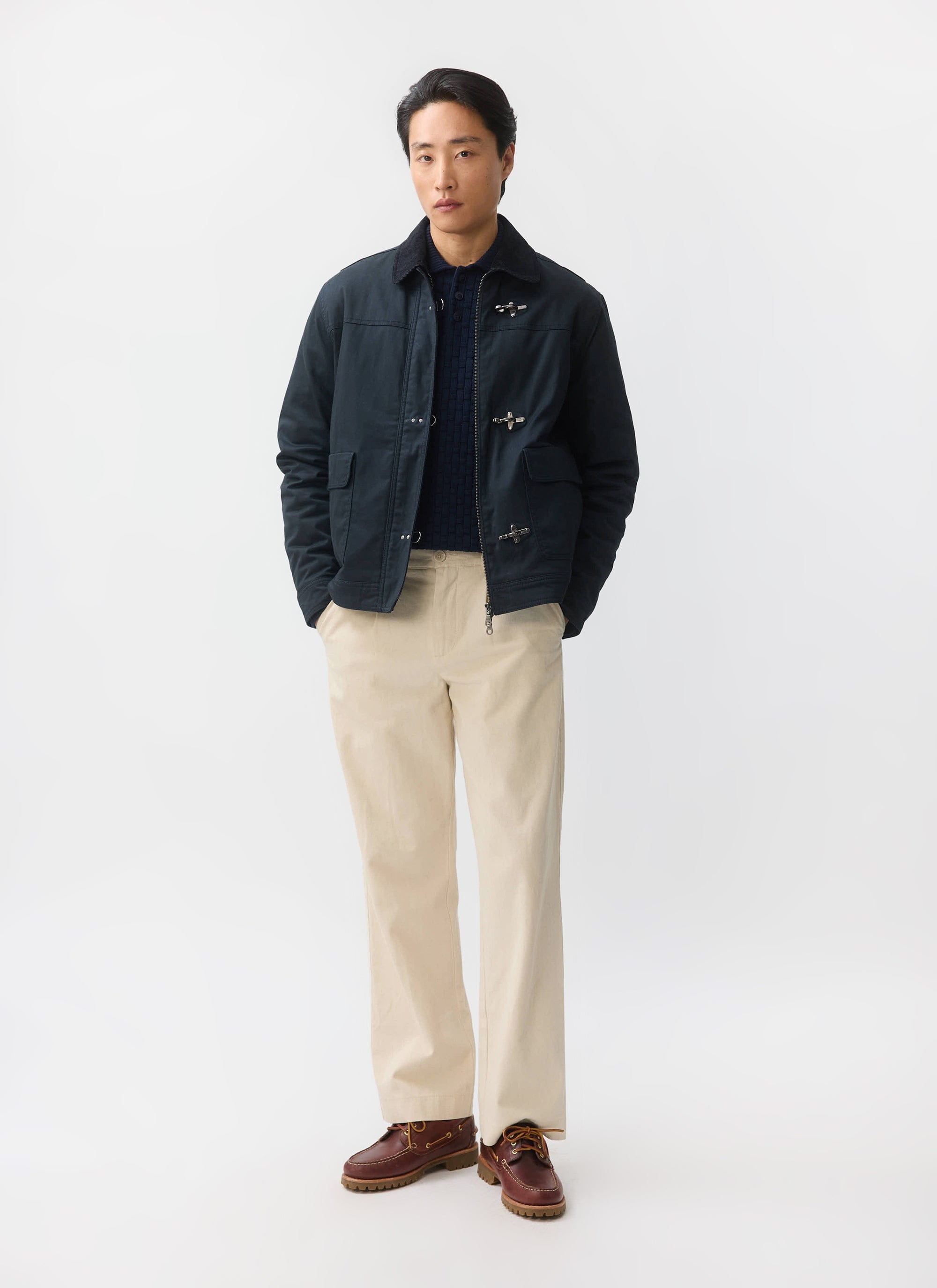 A man models the navy Fontane Wax Jacket from Percival, featuring metal fireman clasps, with beige trousers and boat shoes.