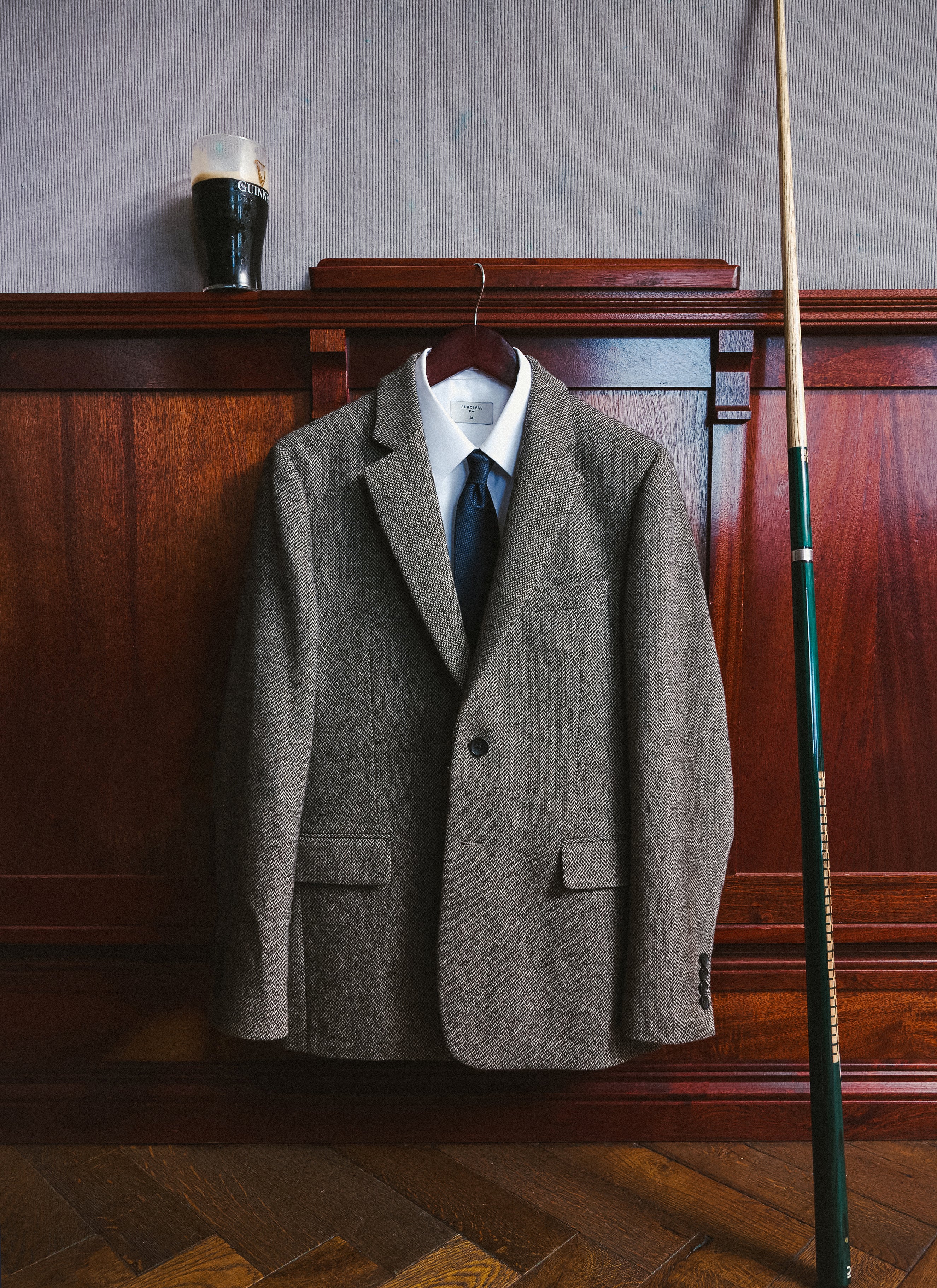 The Percival navy herringbone wool blazer, white shirt and navy tie hanging in a pub next to a pool cue and a pint.