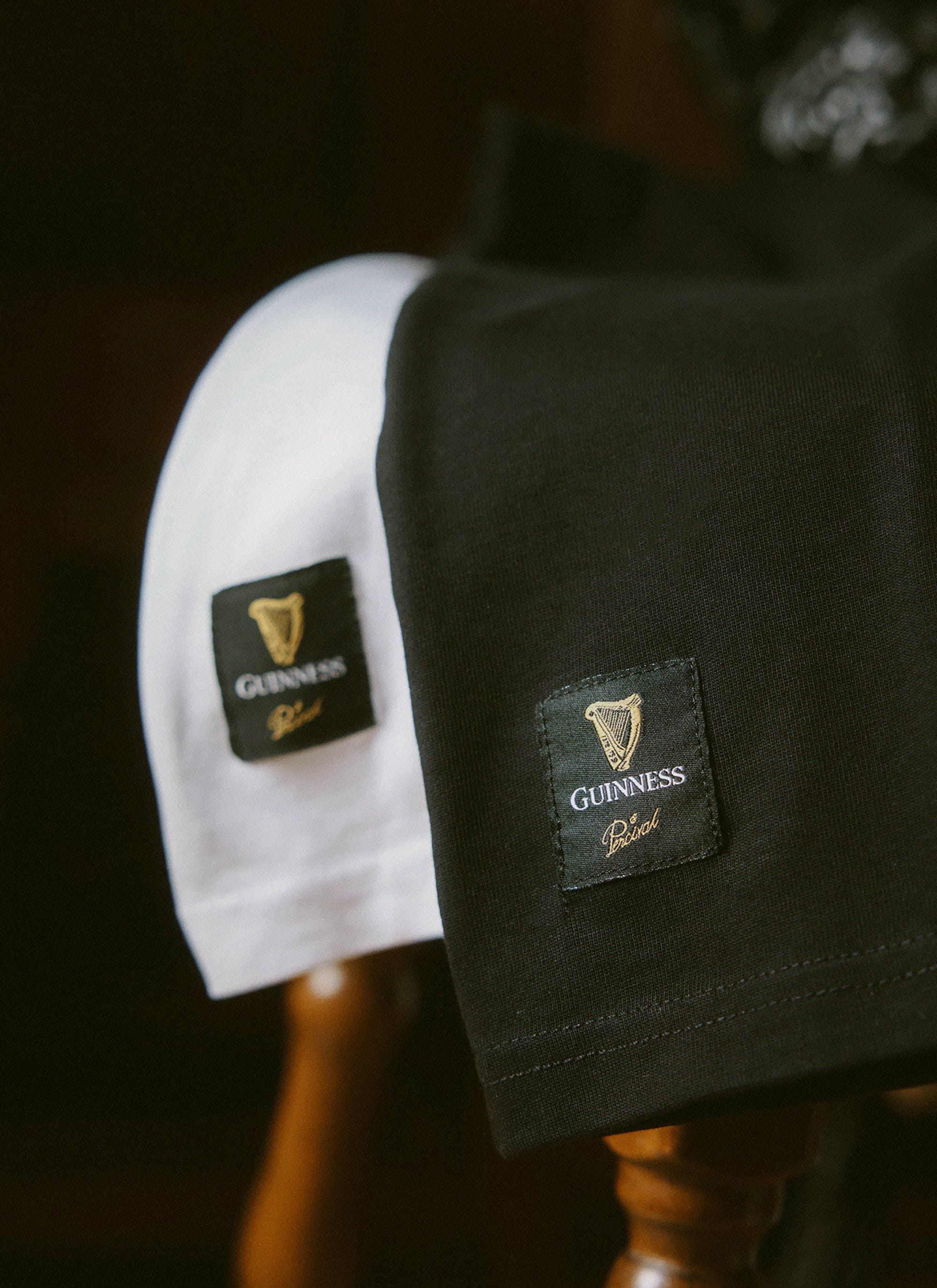 The black and white Percival x Guinness Stout T-shirts draped over a wooden chair, focusing on the woven hem label.
