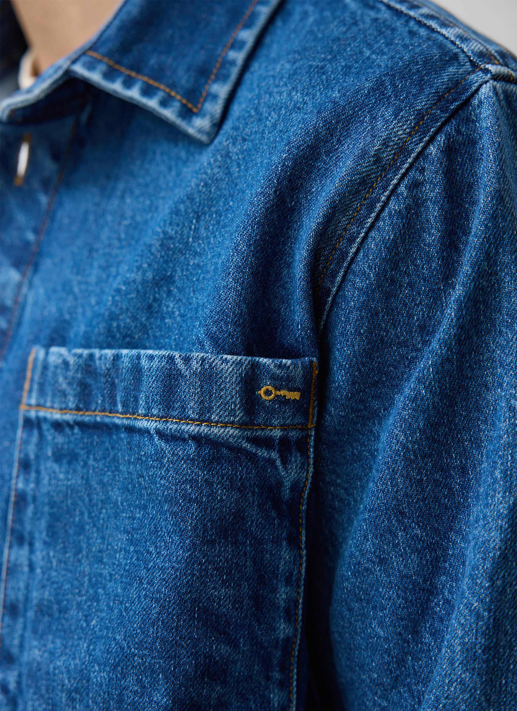 A close-up of the Percival Denim Heavyweight Overshirt in mid-wash blue, showing the chest pocket and collar detail.