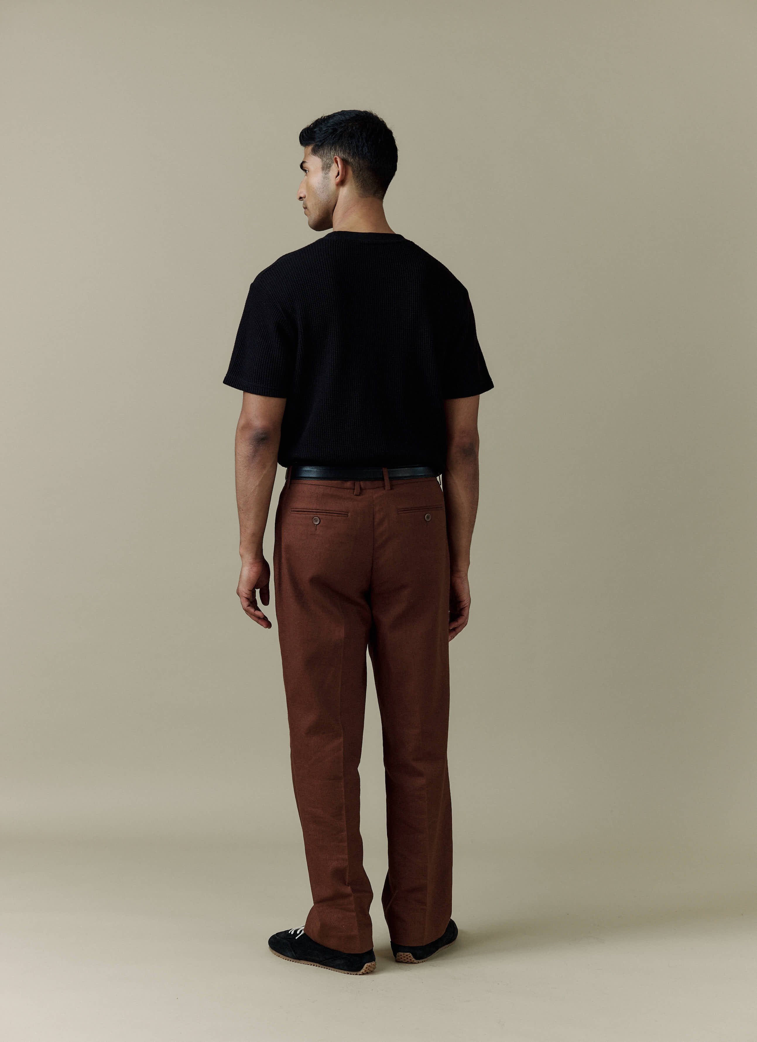 A model seen from the back wearing the Percival Pleated Tailored Linen Trousers in brown, styled with a black t-shirt.