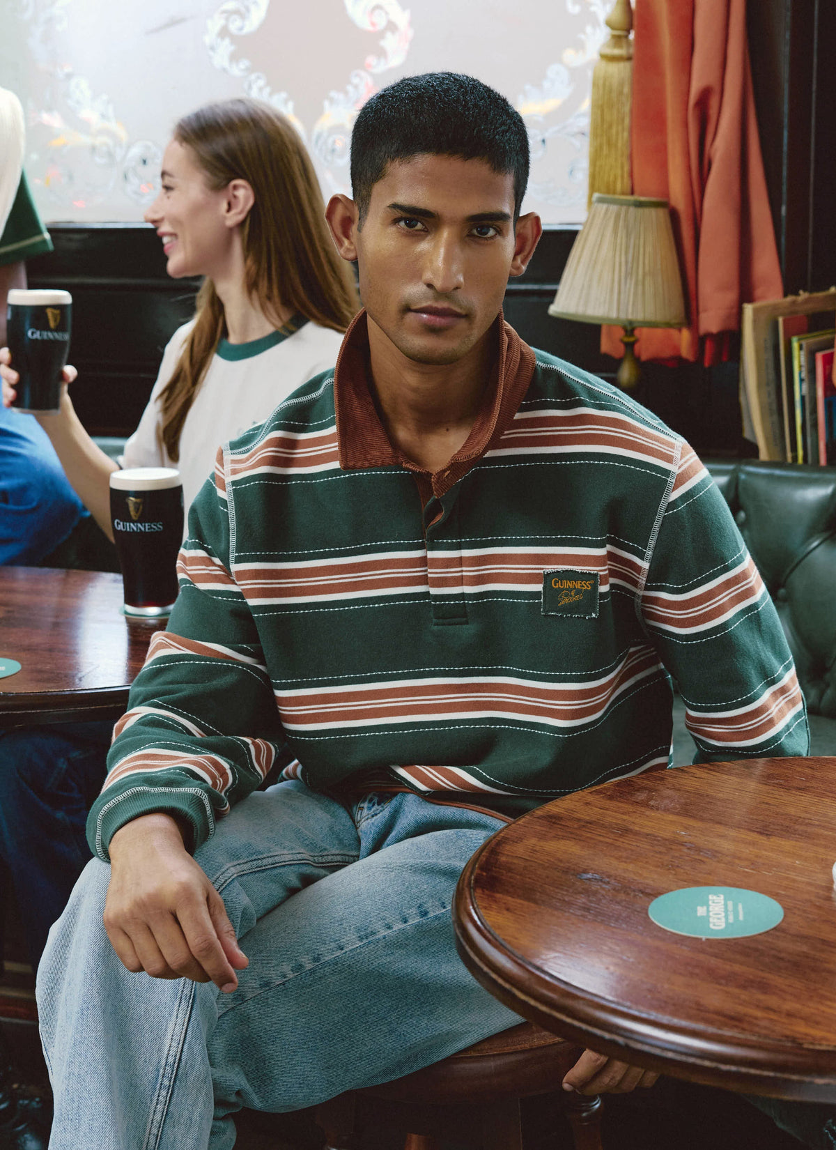 Model wearing the Guinness x Percival striped rugby polo shirt in forest green sitting inside a pub.