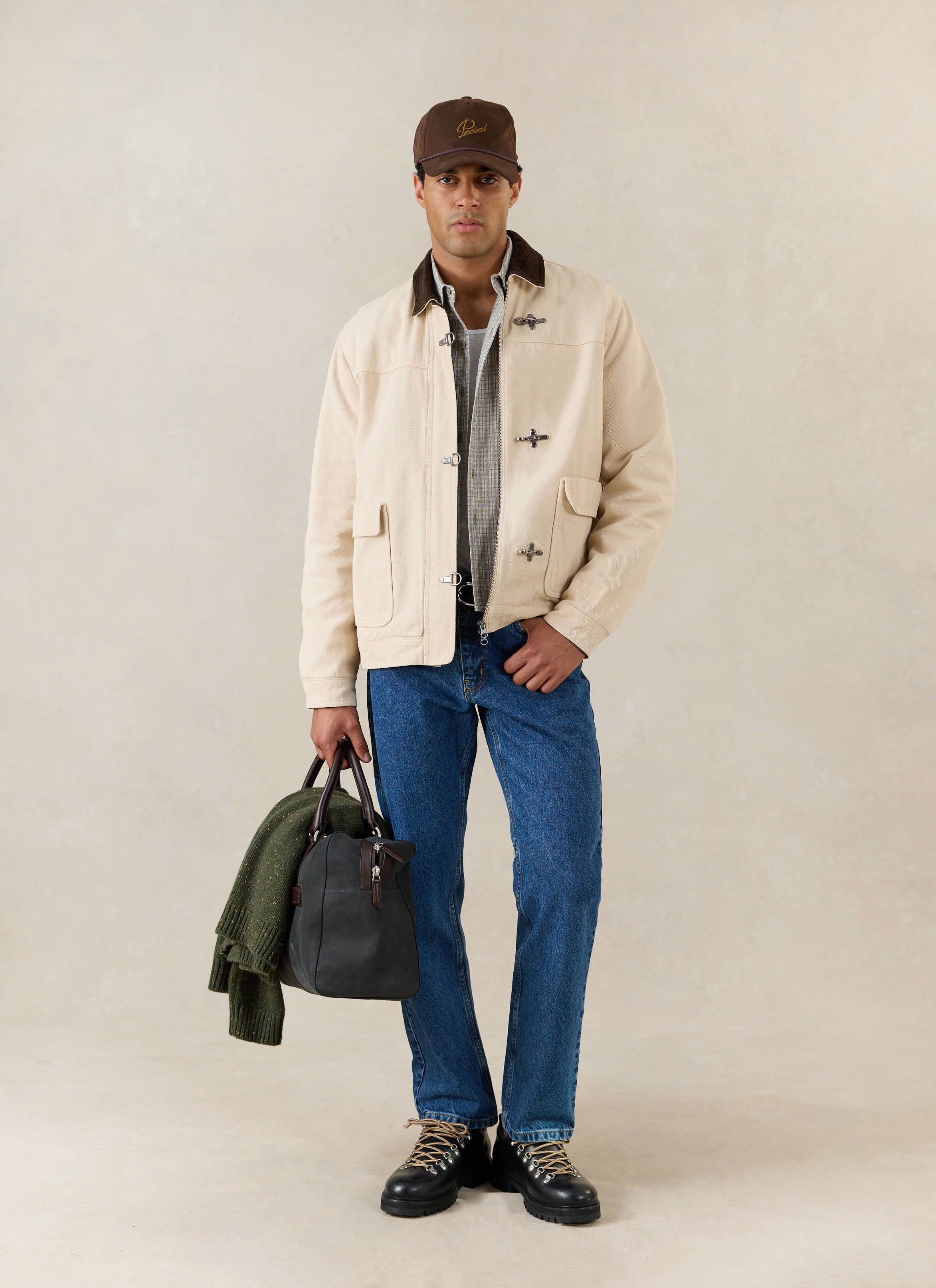 A model wears the cream Percival Fontane cotton fireman clasp jacket, styled with blue jeans, a brown cap, and a holdall.