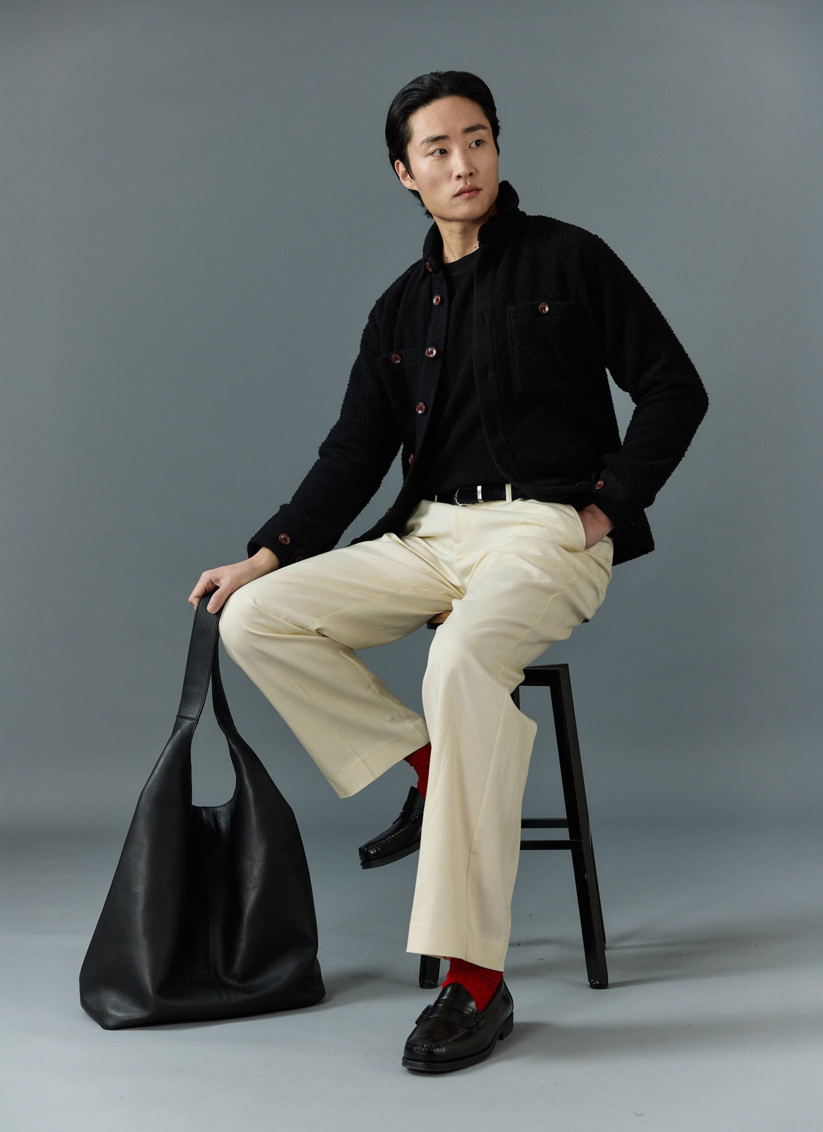 A model sits on a stool wearing the black Percival Blanket Overshirt over a black top with cream trousers and red socks.