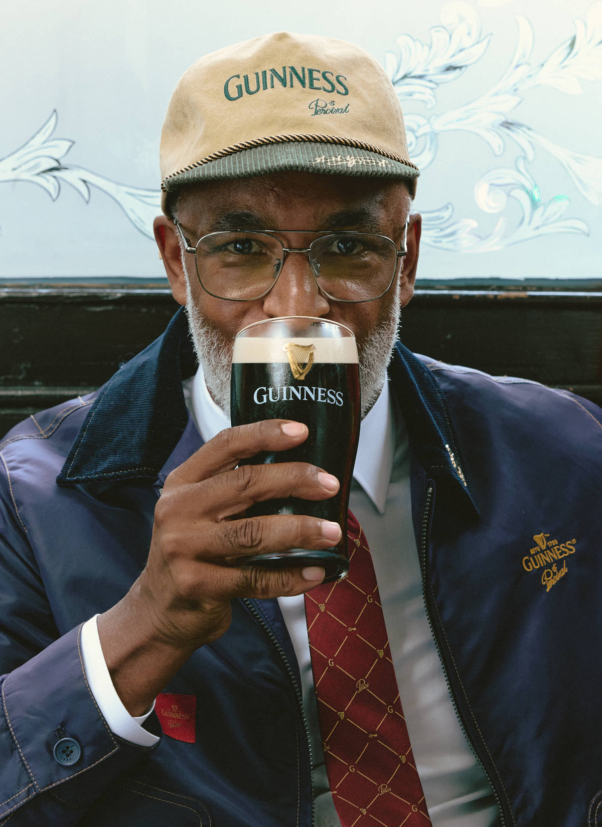 A model drinking a pint of Guinness while wearing the Guinness x Percival beige cap, jacket, and tie.