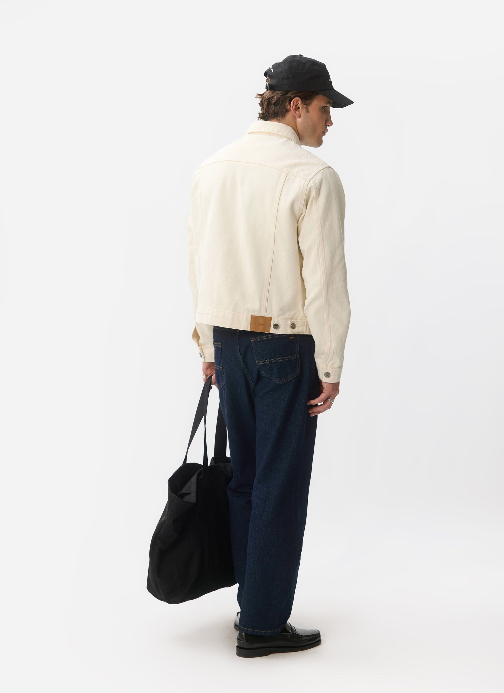A model wears the Percival ecru Western Trucker Jacket, viewed from behind, styled with dark denim and a black tote bag.