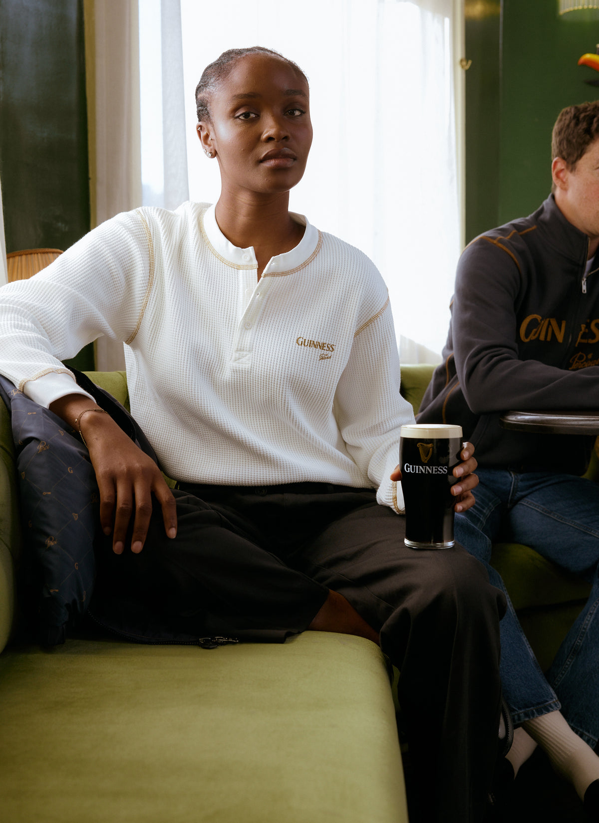 Model sitting on a green sofa wearing a white Guinness x Percival waffle knit top and holding a pint of Guinness.