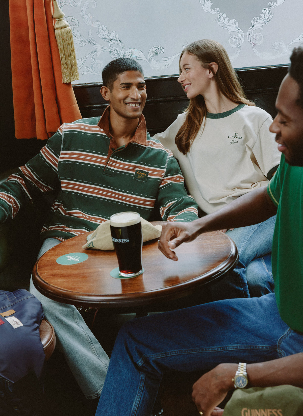 Models relaxing at a pub table wearing the Guinness x Percival collection, with a pint of stout in the foreground.