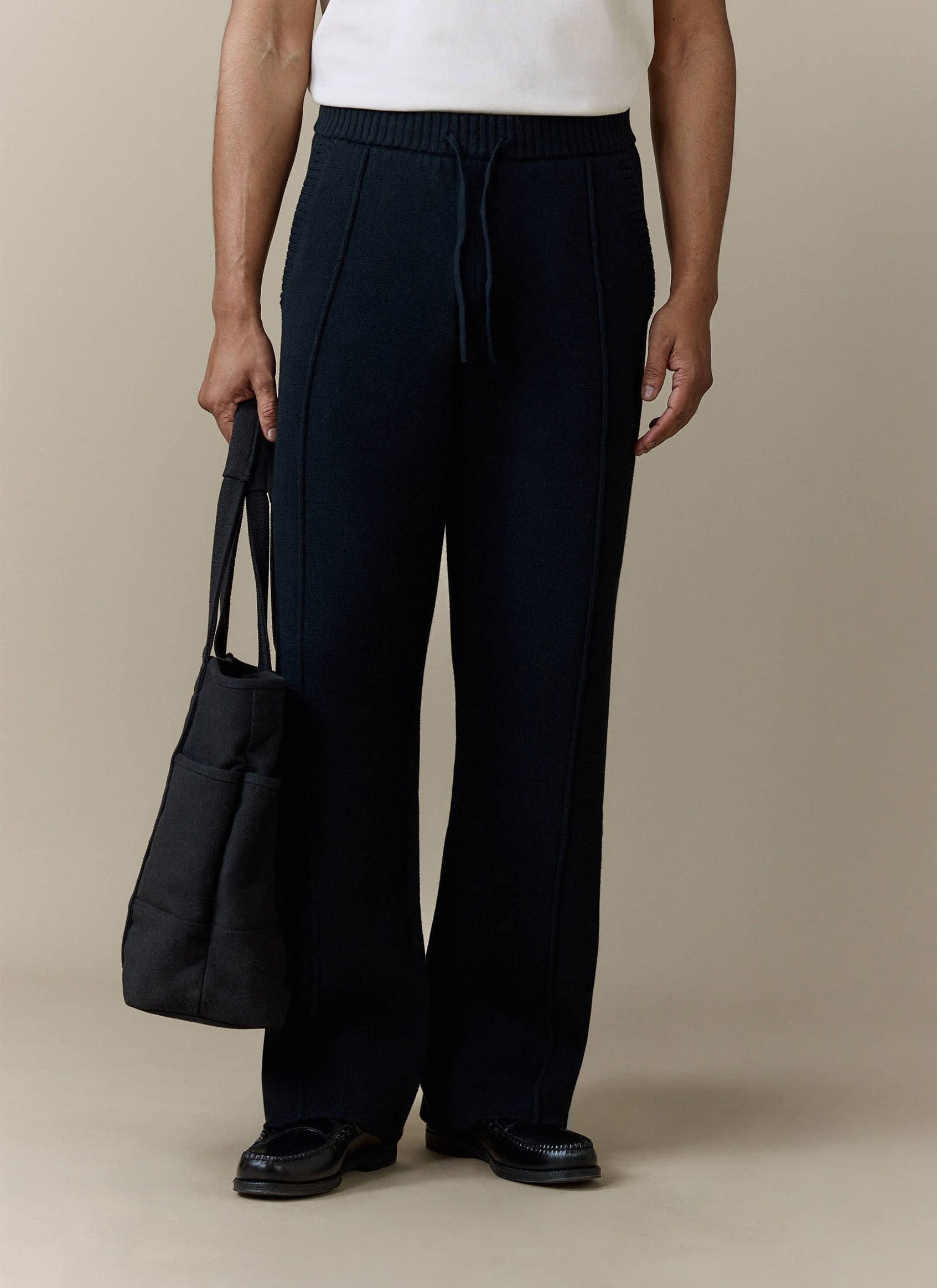A model wears the Percival Chronicle Knitted Straight Leg Trackpant in black, styled with loafers and holding a tote bag.