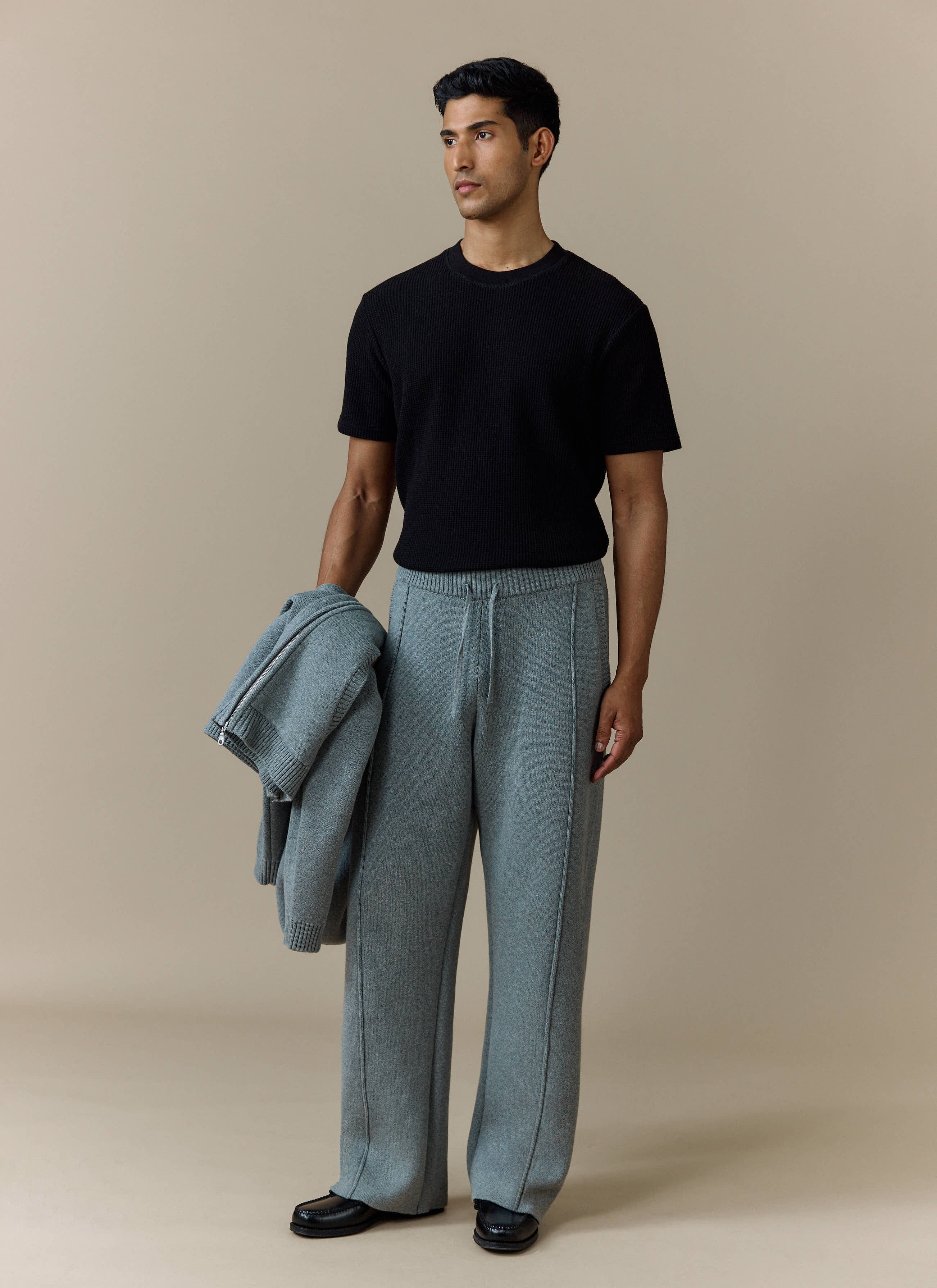 A model wears the Percival Chronicle Knitted Straight Leg Trackpant in grey with a black tee, holding a matching grey jacket.