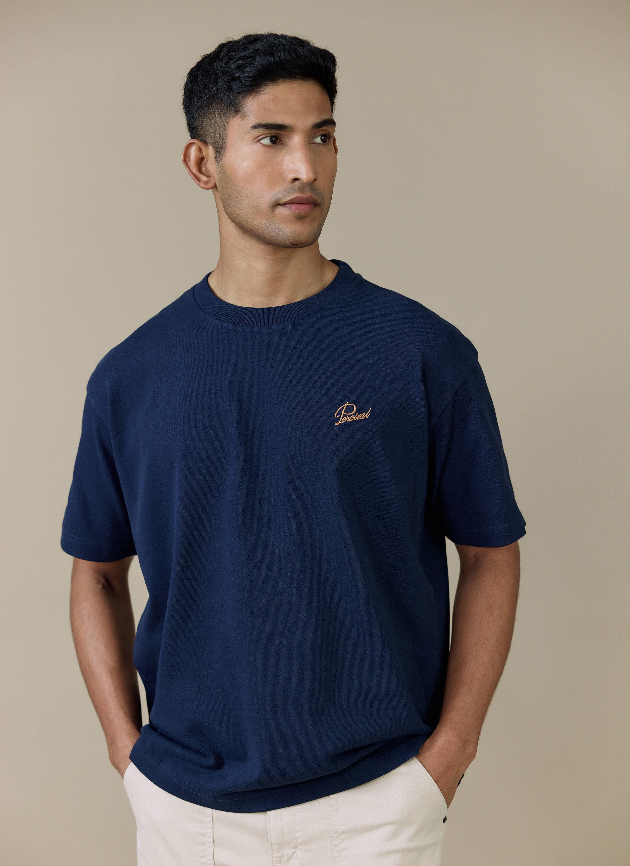 A model wearing the navy Percival Perci Script T Shirt in organic cotton, featuring contrast chest script embroidery.