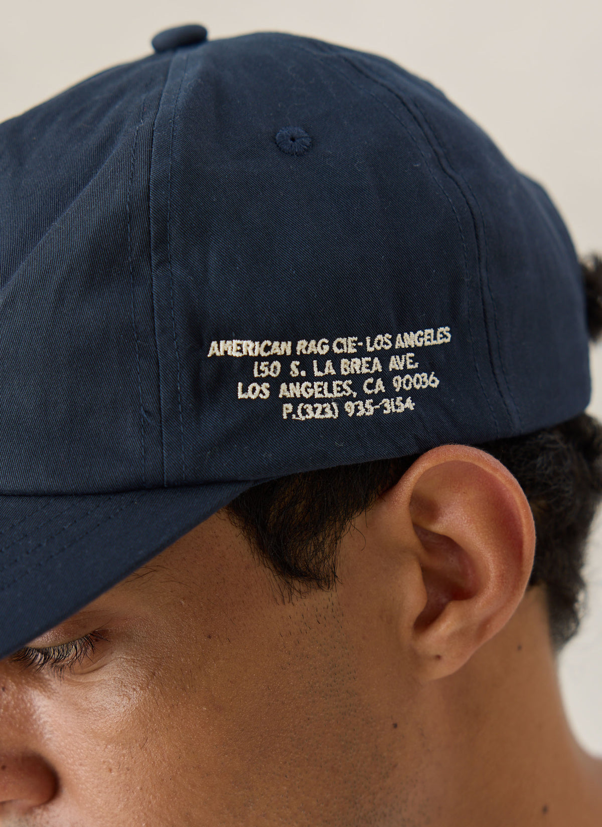 A close-up of the navy American Rag x Percival cap showing white embroidered address lettering on the side.