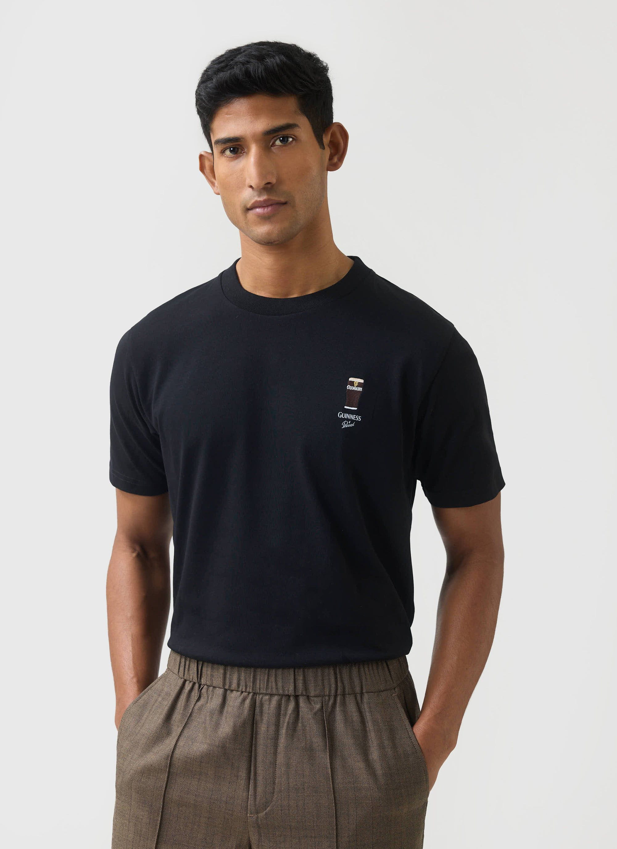 A man models the black Stout T-shirt from the Percival x Guinness collaboration, paired with brown patterned trousers.