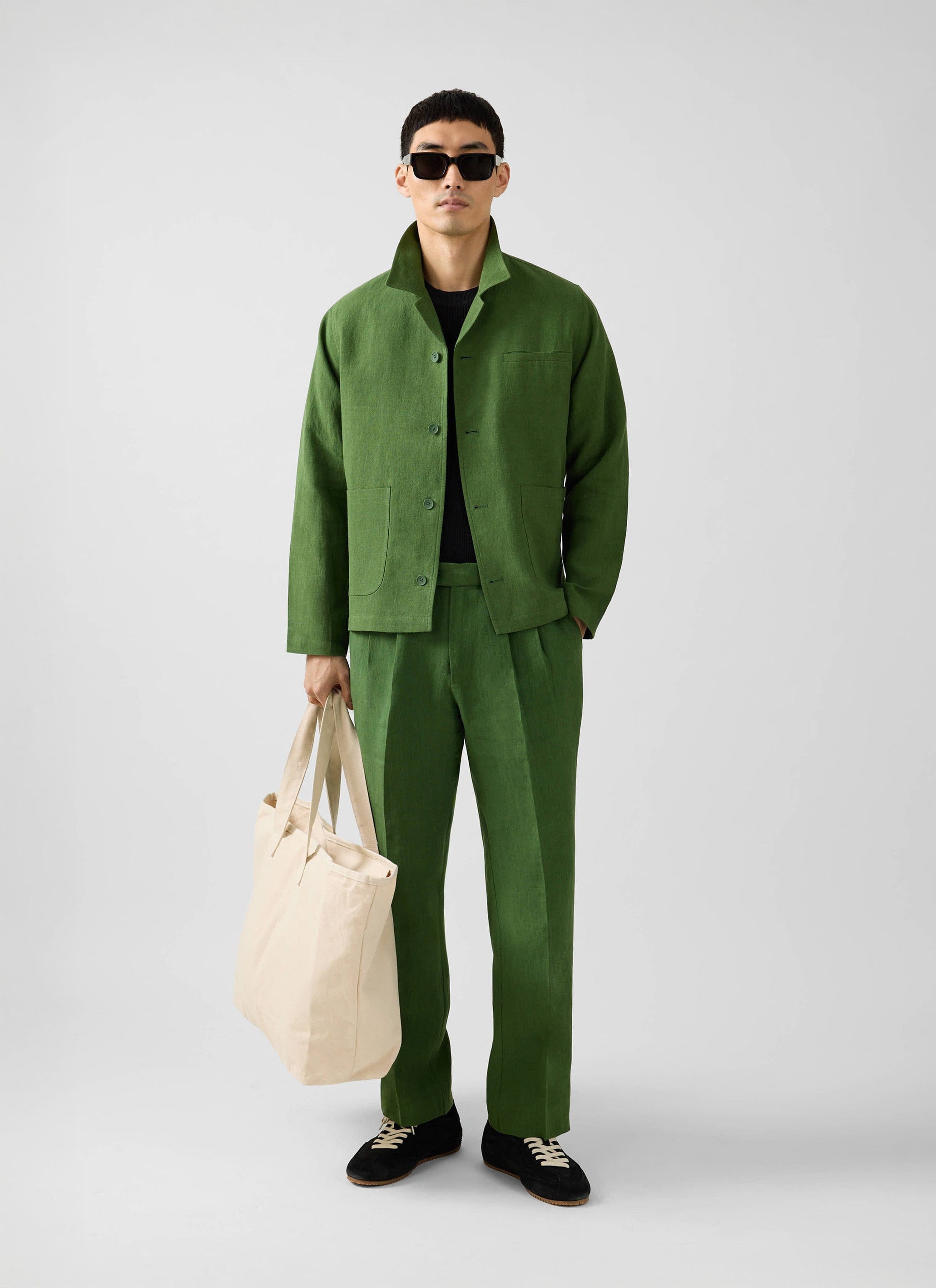A man models the Percival forest green linen Overshirt and matching trousers, wearing sunglasses and holding a tote bag.