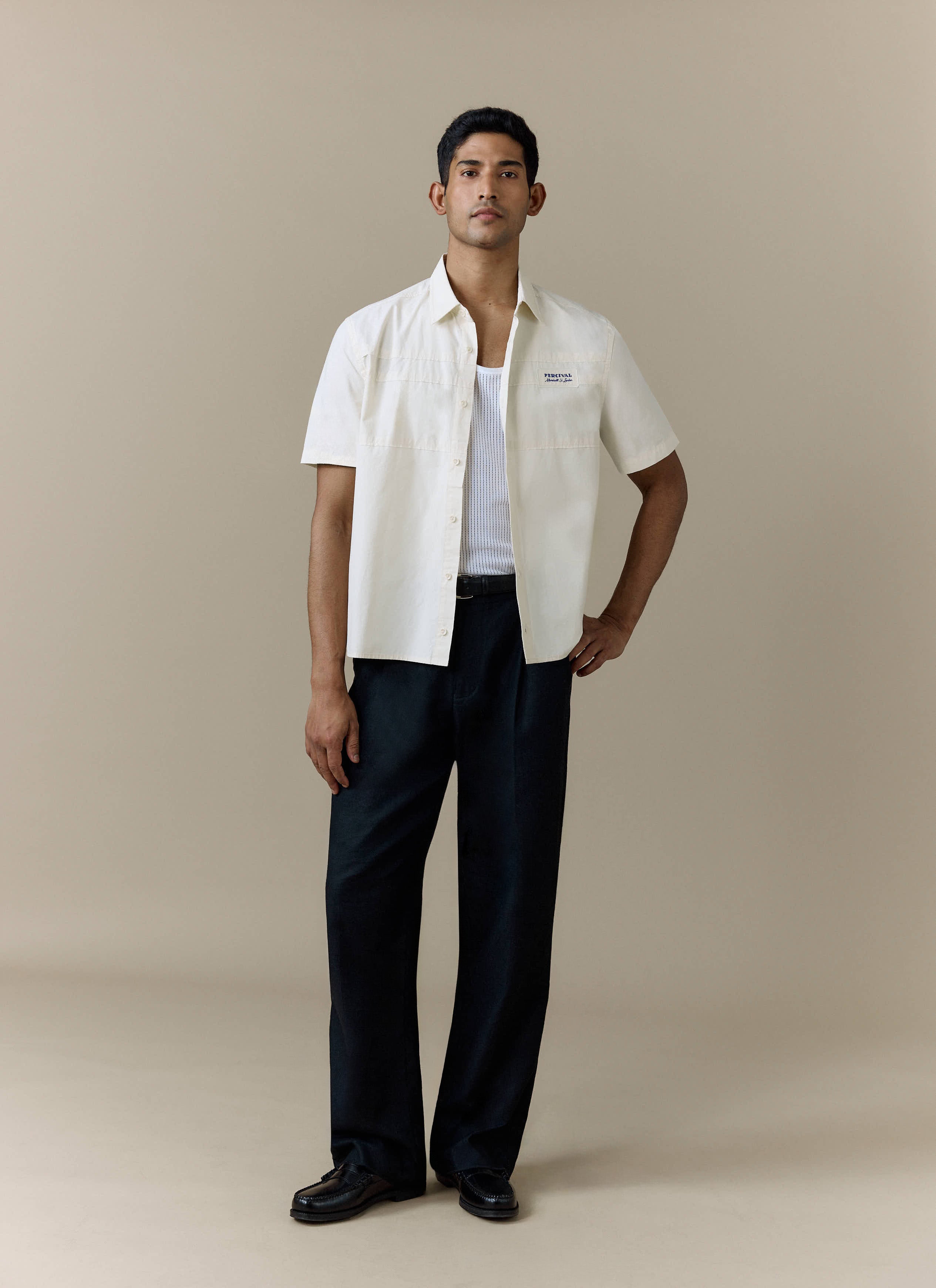 A model wears the Percival Harlow Boxy Shirt in ecru, styled open over a white vest with wide-leg black trousers.