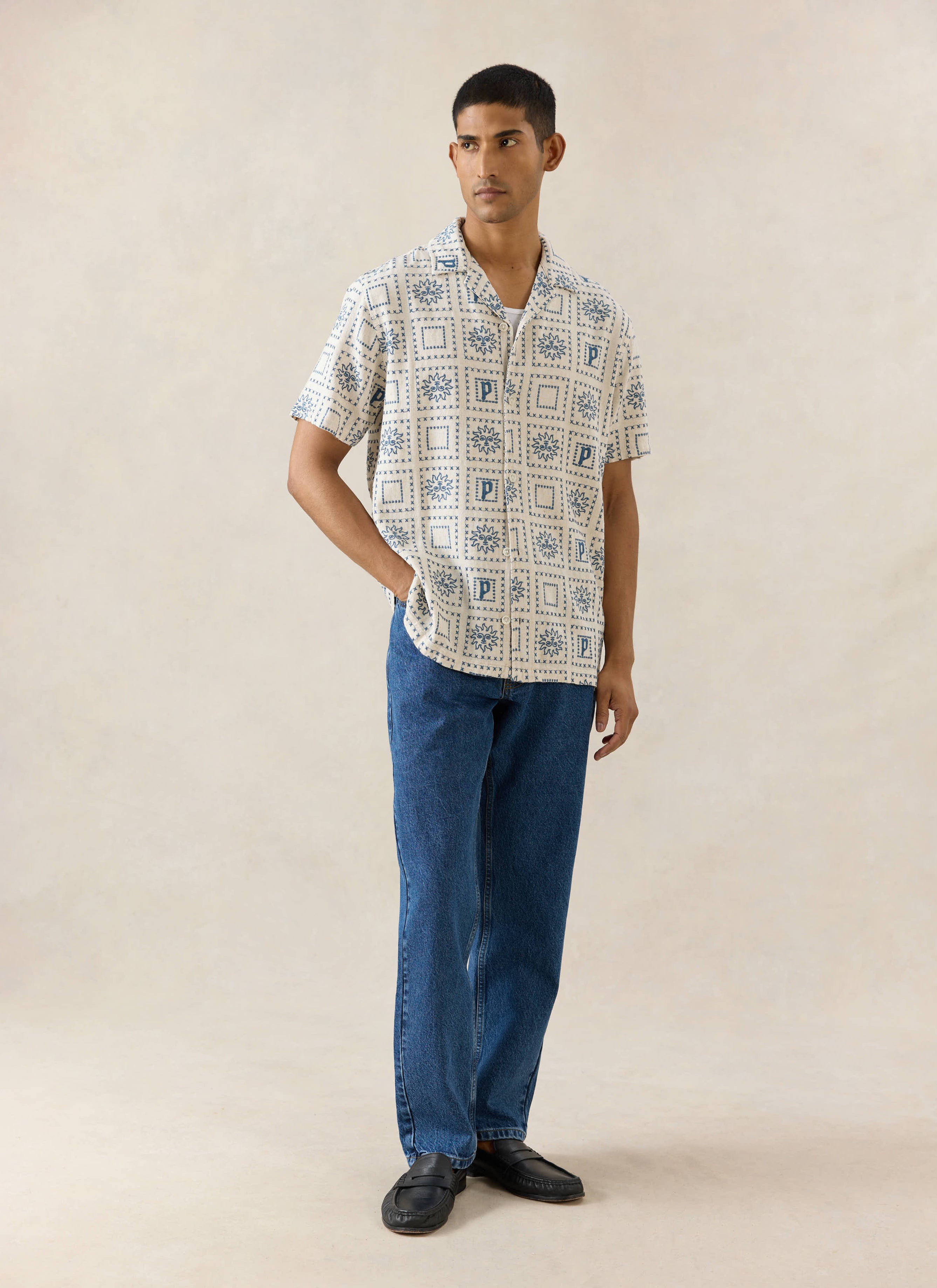 The Percival Solis Heaton Cuban shirt in cream linen blend, featuring a blue sun pattern, styled with denim jeans.