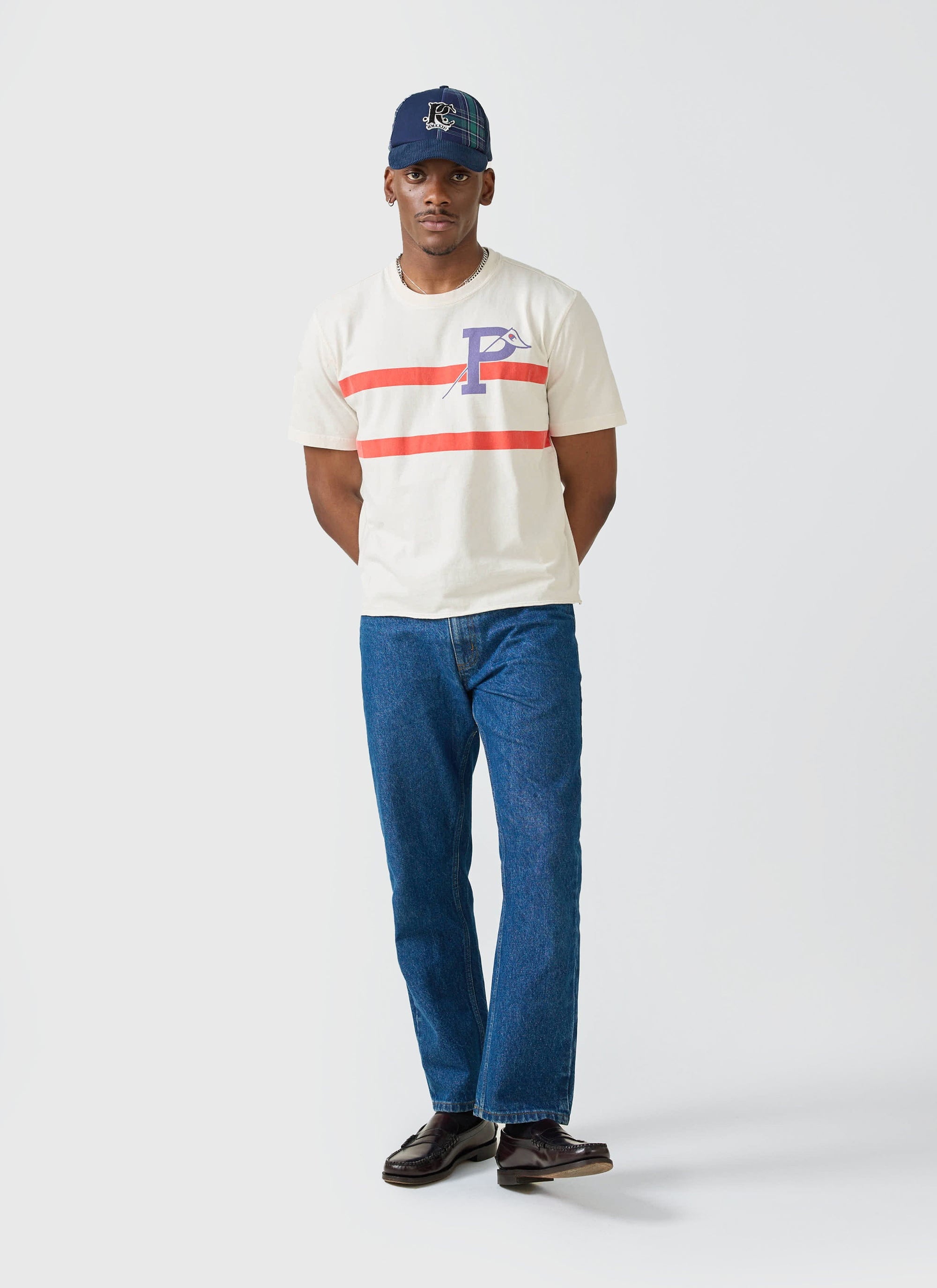 A model wears the Percival x Champion Flag Stripe T-shirt in ecru, styled with dark wash jeans and a blue cap.