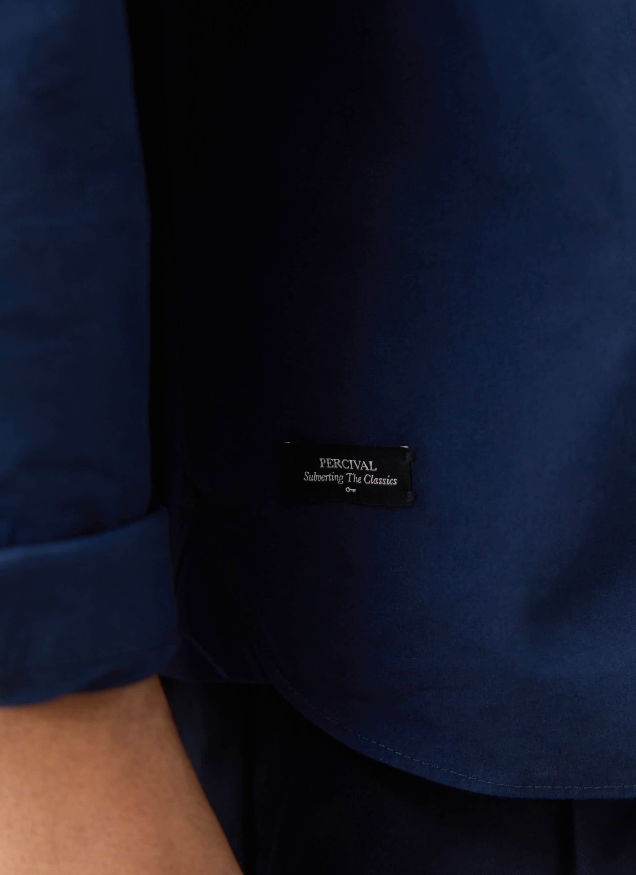 Close-up of the navy cotton Percival Dali Relaxed Poplin Shirt, featuring the 'Subverting The Classics' woven hem label.