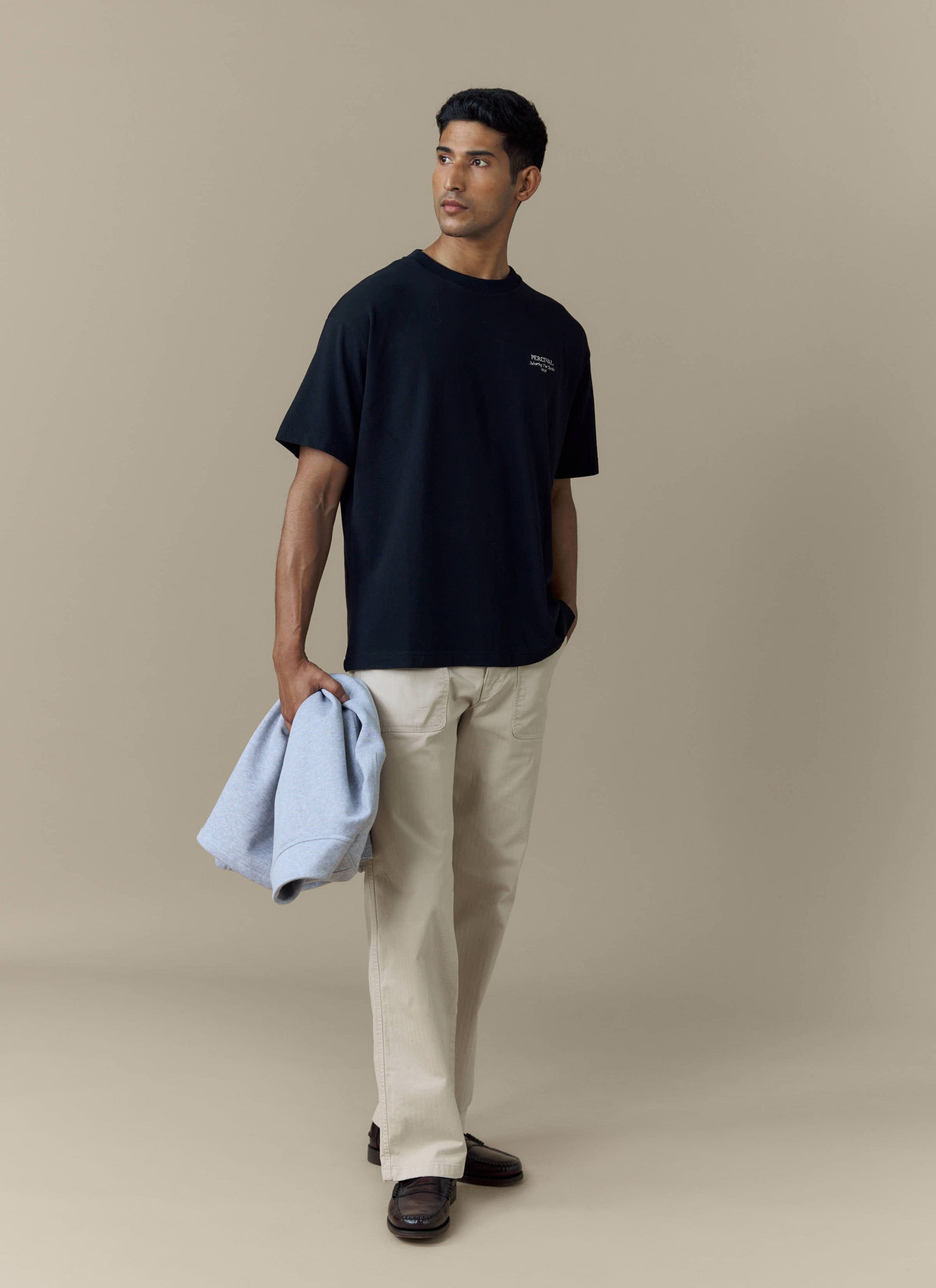 A model wears the Percival Fruit Farm Oversized T-Shirt in black, styled with cream trousers and holding a blue sweatshirt.