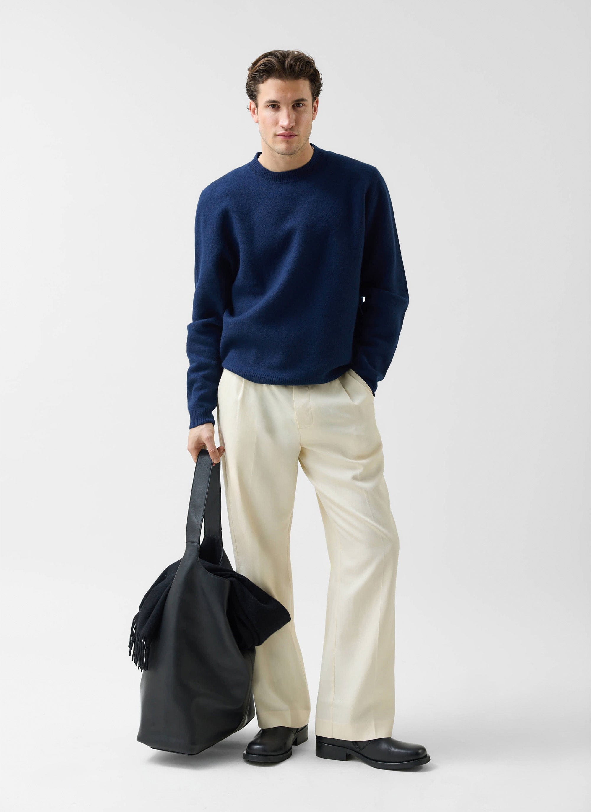 A man models the navy Felted Merino Wool Jumper from Percival with cream trousers and a black tote bag.