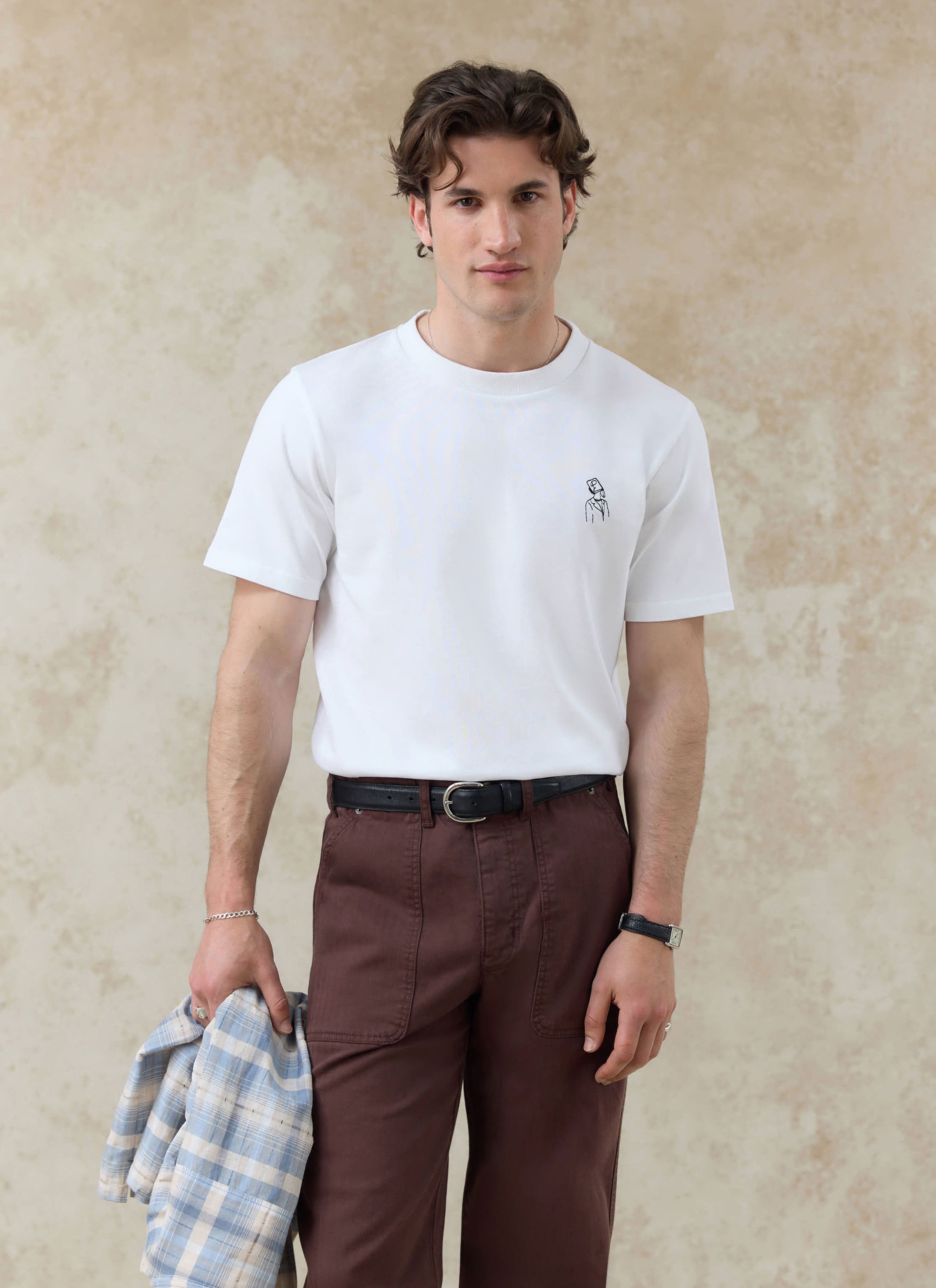 White Percival Robinson organic cotton t-shirt with black chest embroidery, styled with brown trousers on a model.