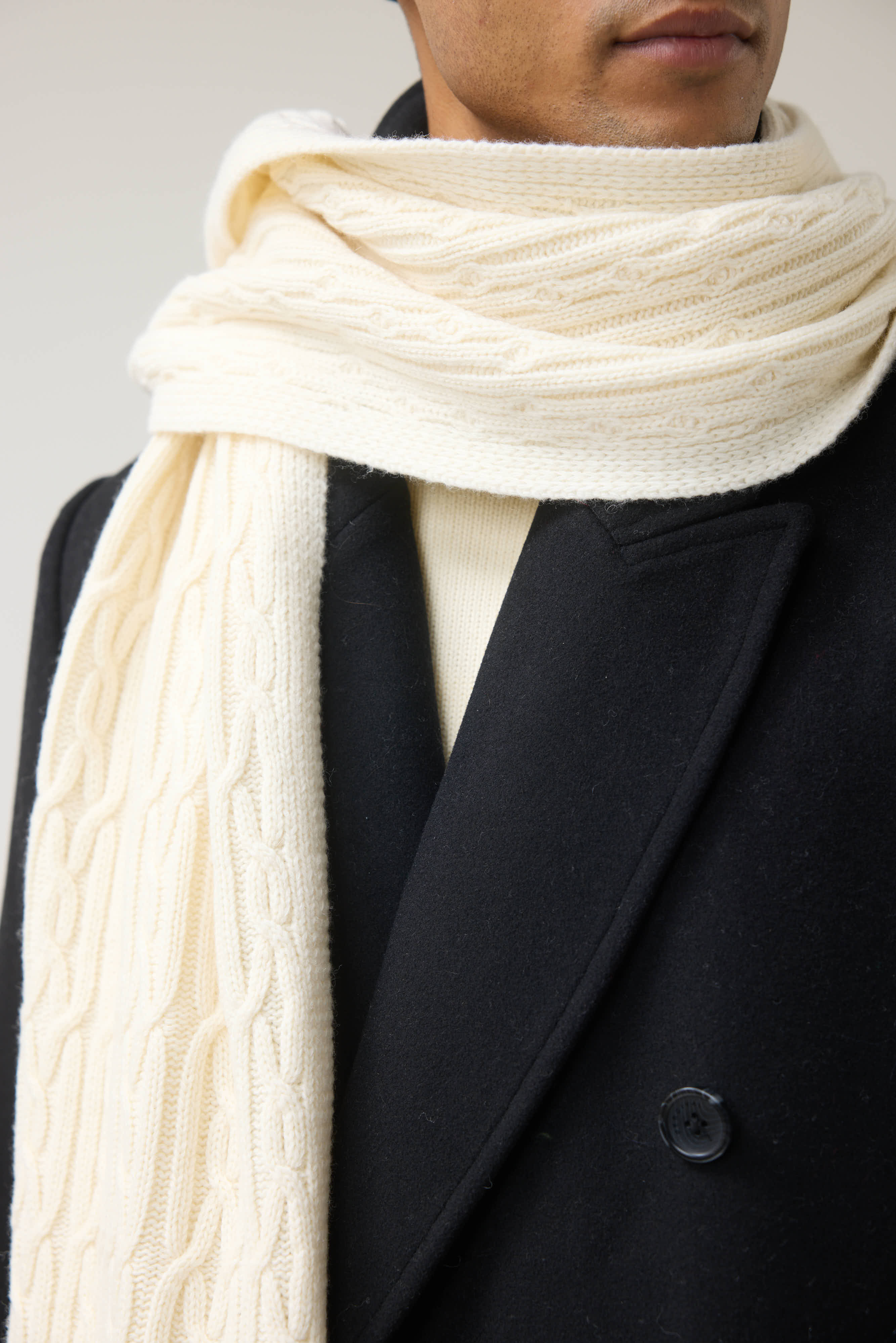 A close-up of a man wearing the ecru wool Melville Cable Knit Scarf from Percival over a dark coat.
