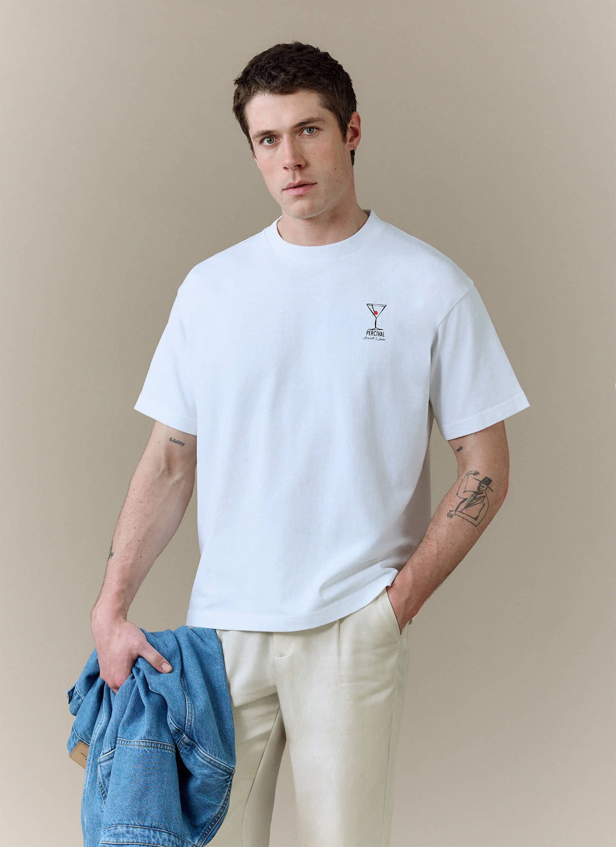 A model wears the white Perci and Rossi Martini oversized t-shirt from Percival with cream trousers and holds a denim jacket.