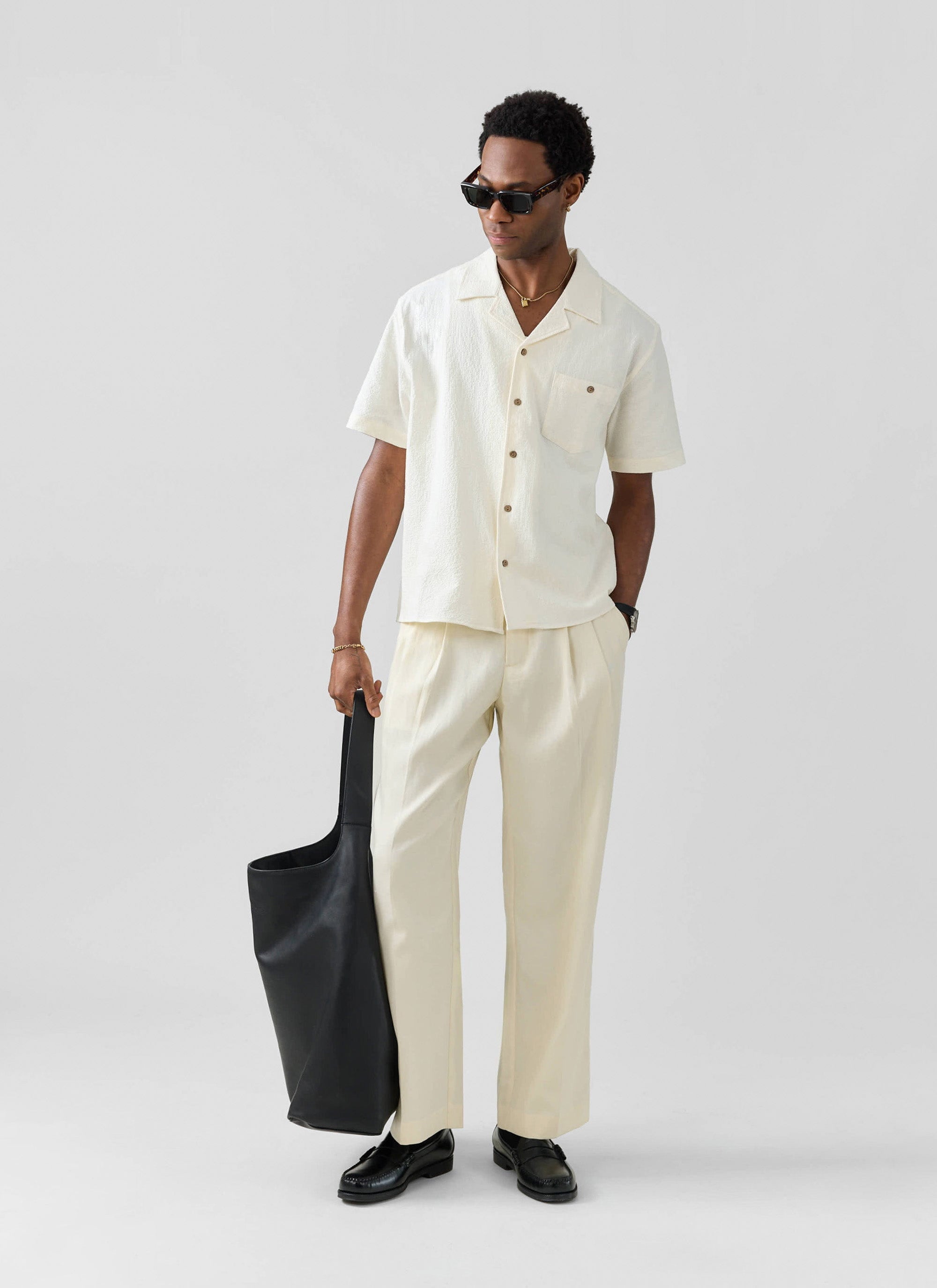 A model wears the cream Pleated Tailored Trousers from Percival with a matching shirt, sunglasses, and a black tote bag.